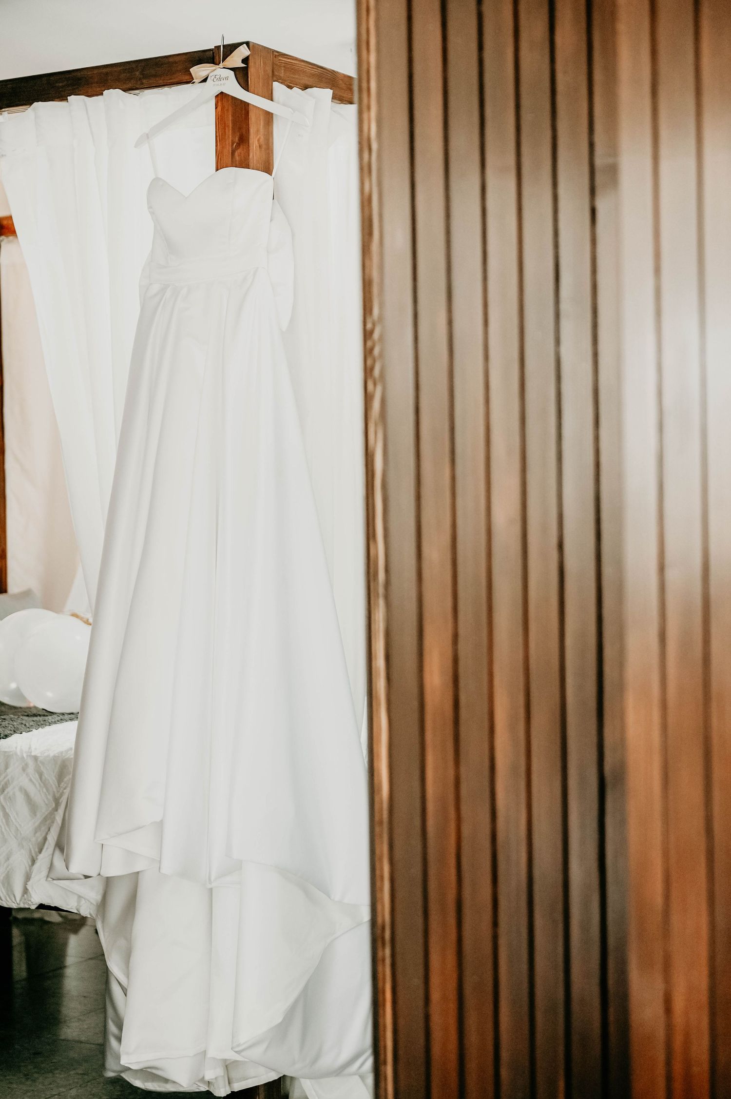 A white wedding gown with a sweetheart neckline hangs against a wooden paneled wall in warm lighting.
