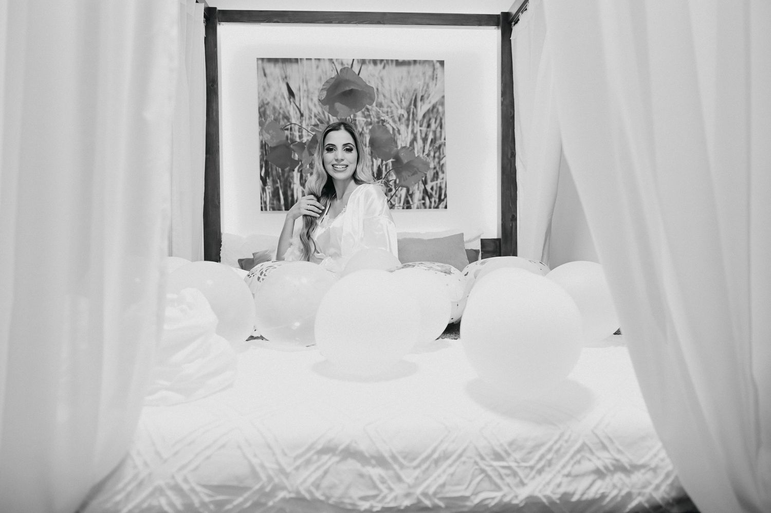 Black and white photo of someone in bed taking a selfie in the mirror, with white curtains and bedding visible.