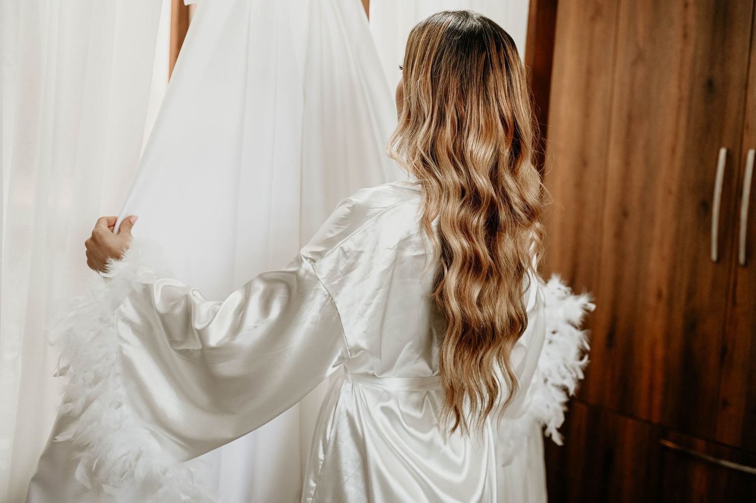 Long wavy hair cascades down the back of someone wearing a flowy white robe while holding a wedding dress.