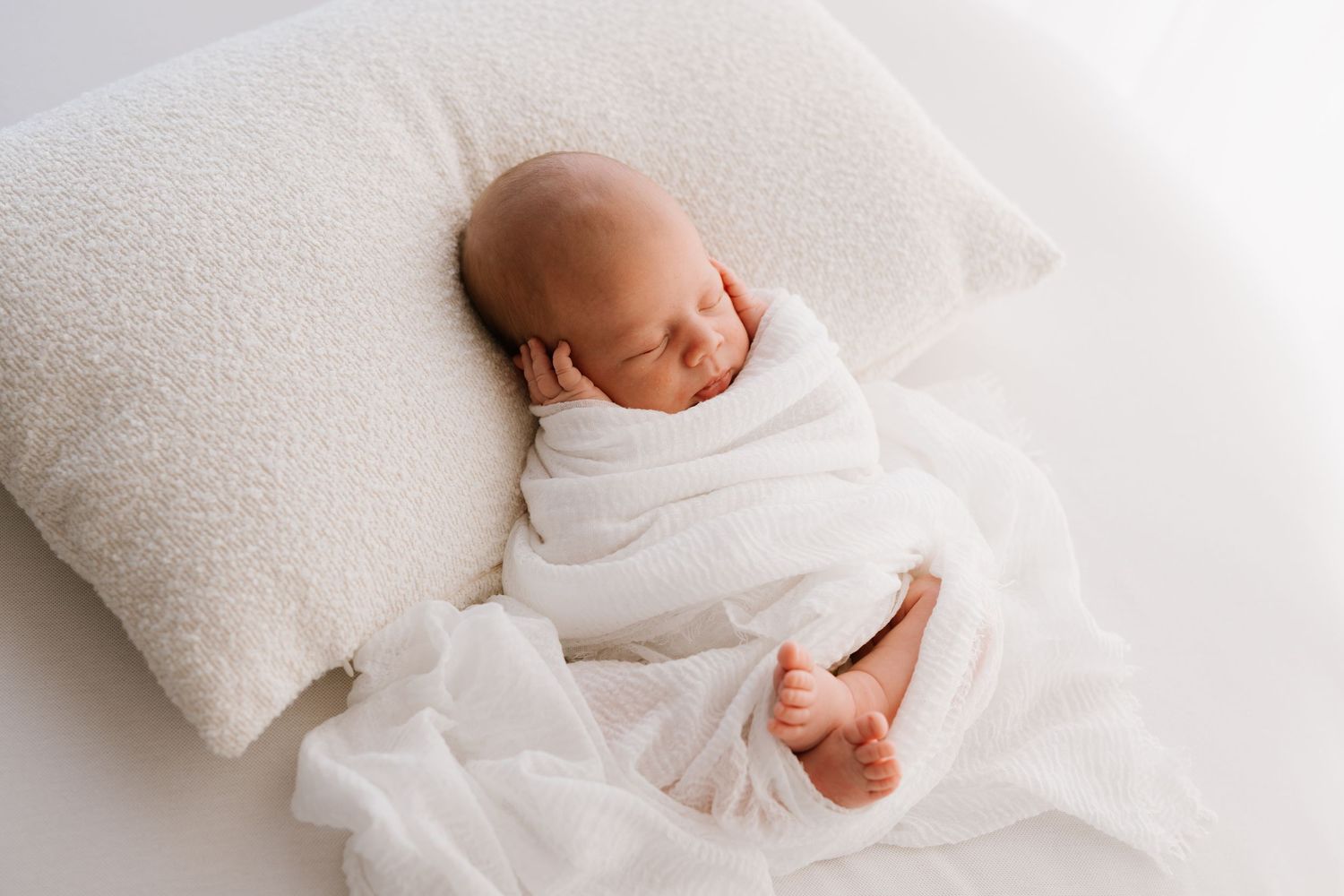 Sleeping newborn baby swaddled in white wrap resting on fluffy pillow in bright studio setting.