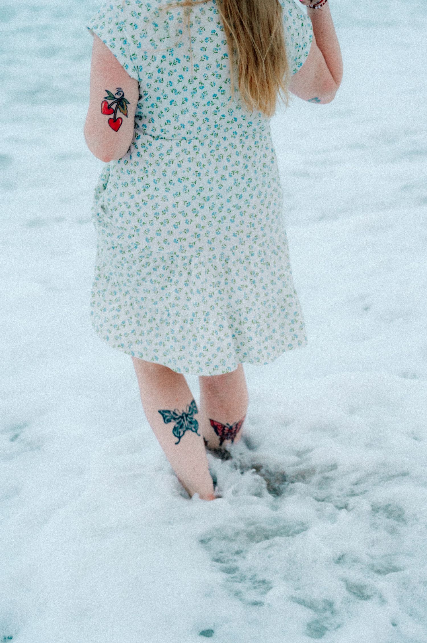 Someone in a floral dress stands barefoot in snow showing colorful butterfly and rose tattoos on their legs and arms.