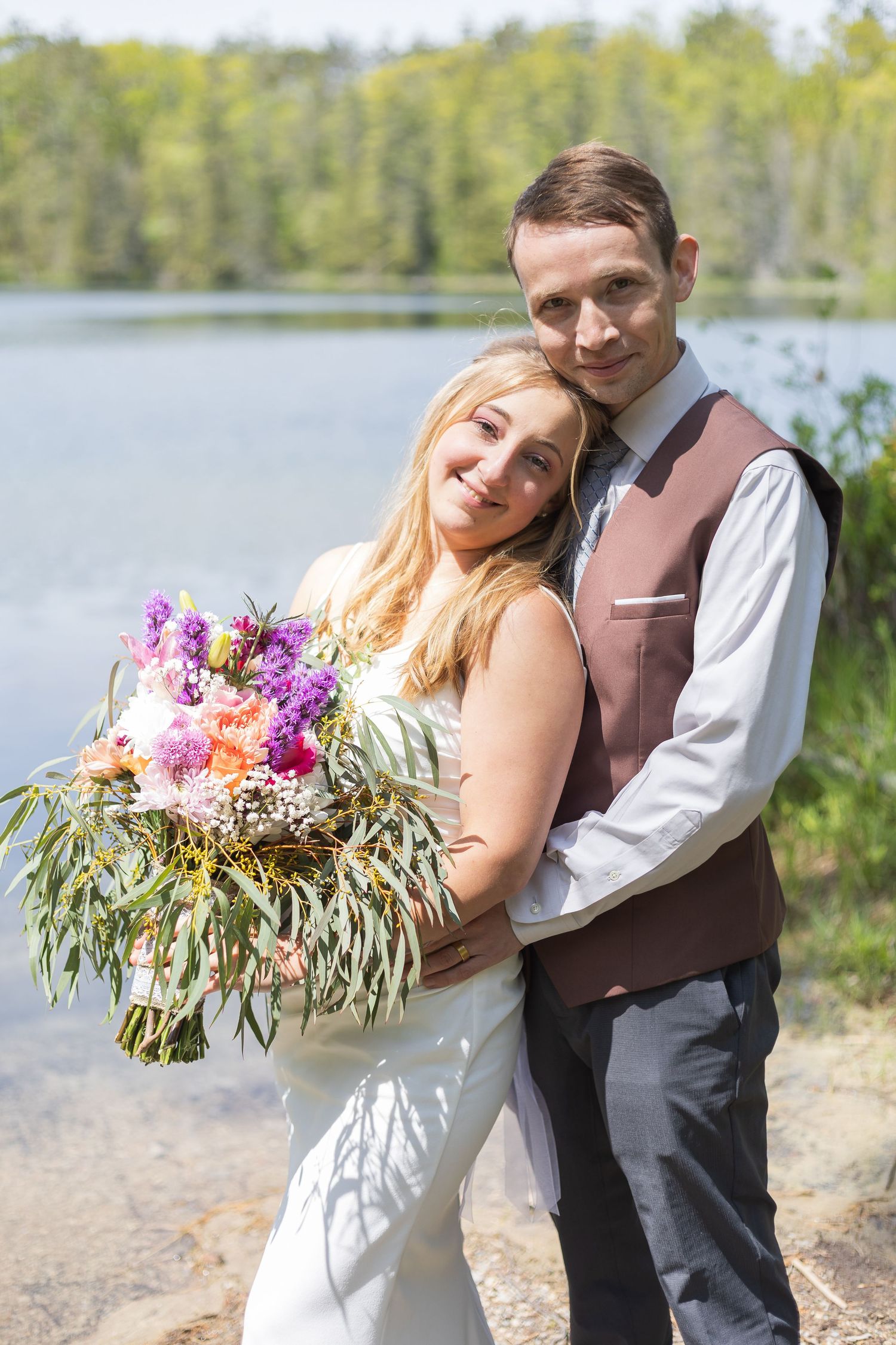 Wedding couple embracing by a lake with a colorful bouquet of flowers.