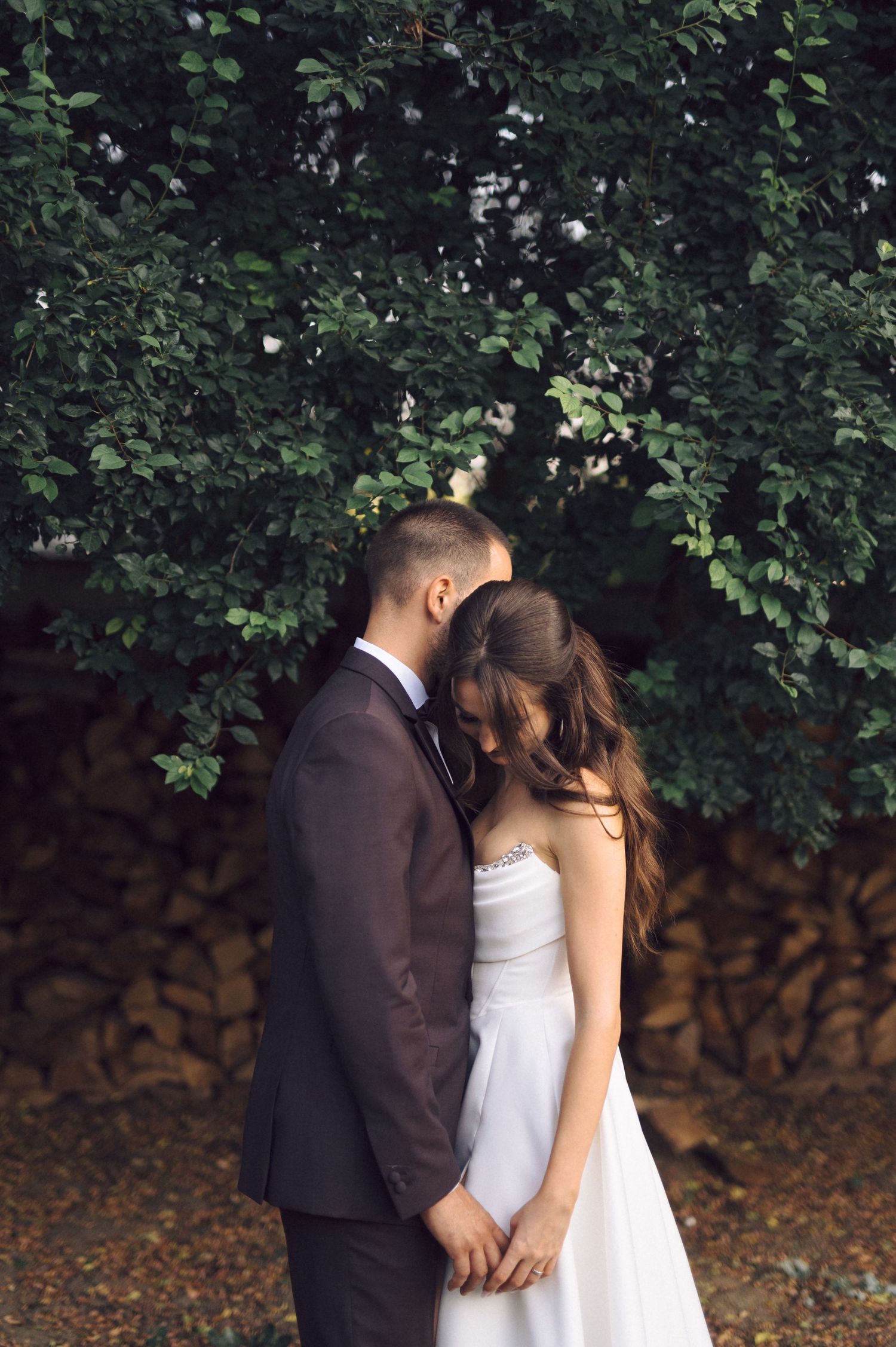 A romantic newlywed couple shares an intimate moment together among green foliage in an outdoor setting.