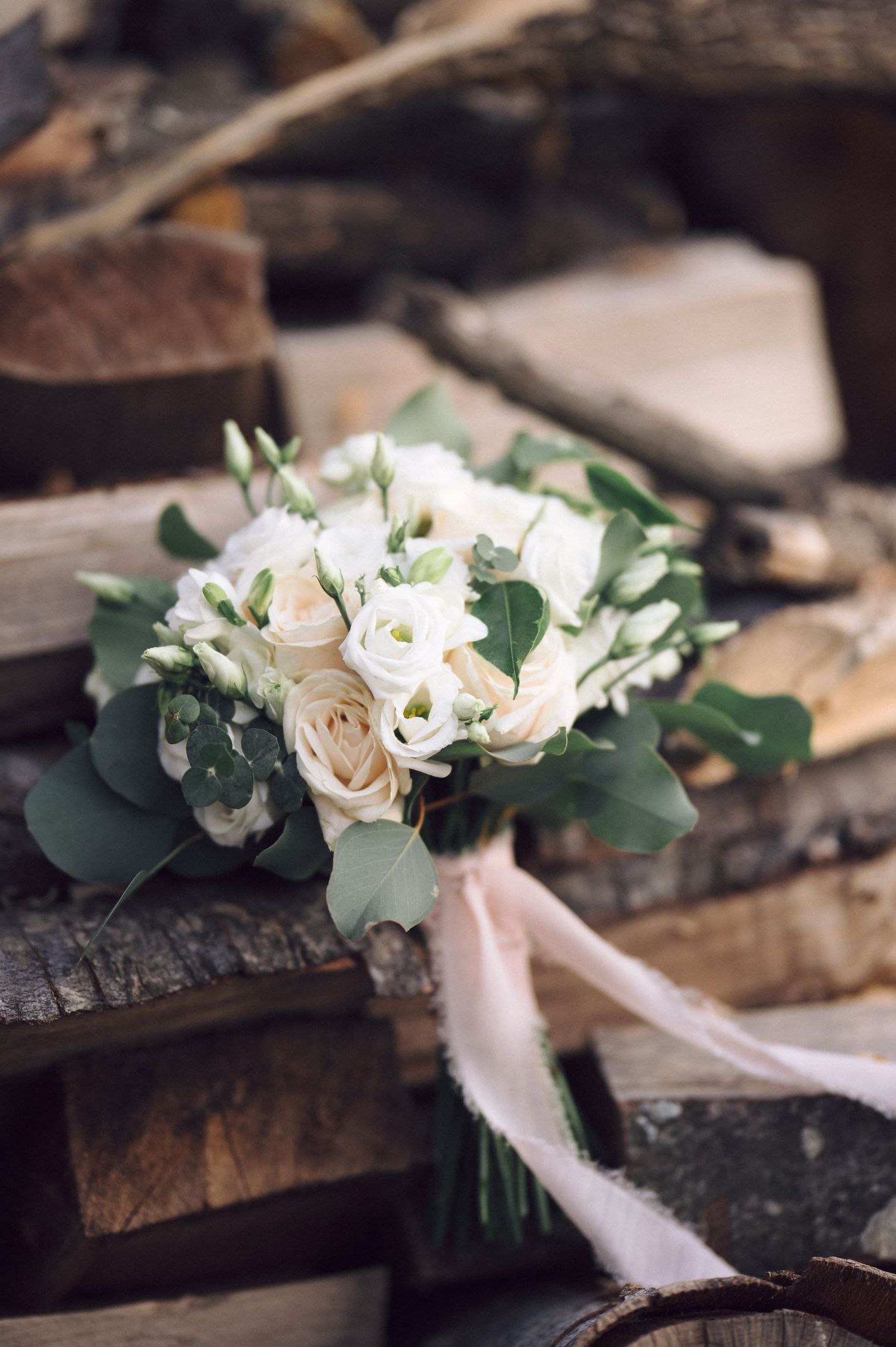 Elegant white rose and eucalyptus wedding bouquet with silk ribbons resting on rustic wooden surface.