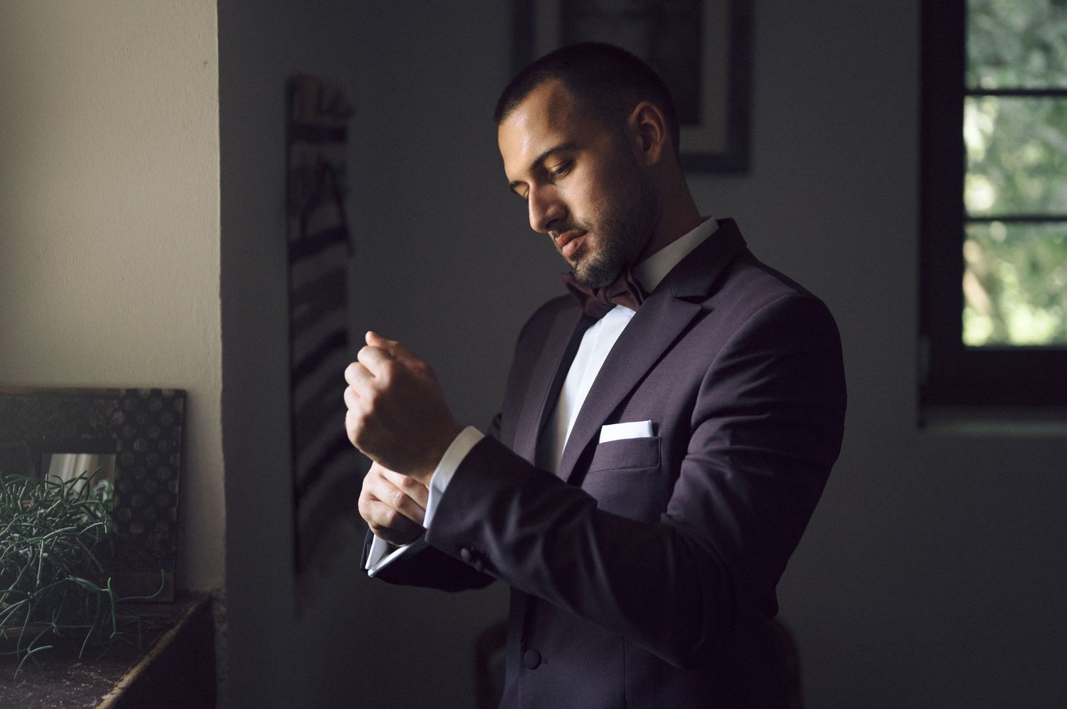 A groom in a dark suit adjusts his tie while standing in a dimly lit room by a window.