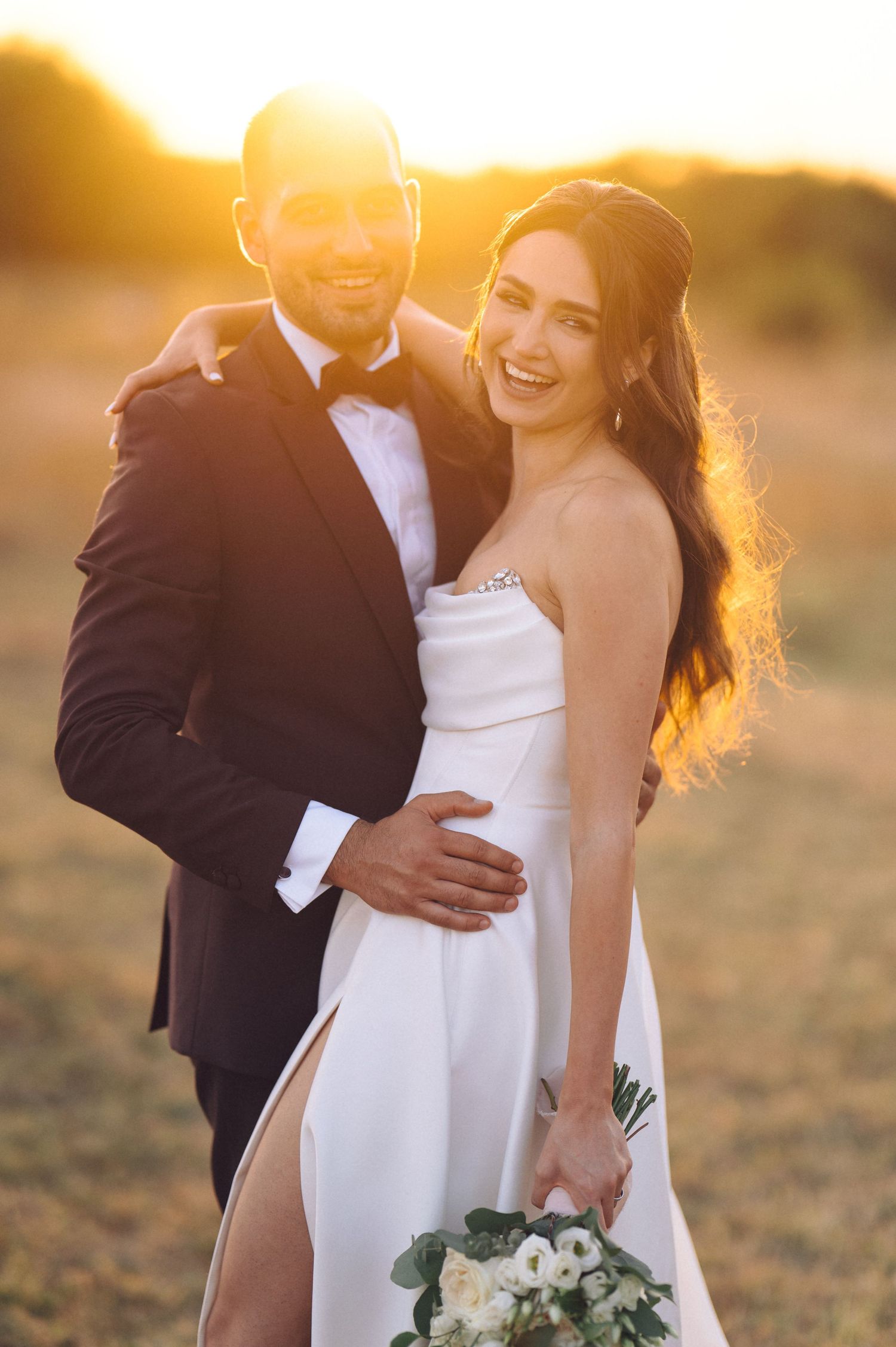 Golden sunset light illuminates couple in wedding attire embracing outdoors with white bouquet of flowers.