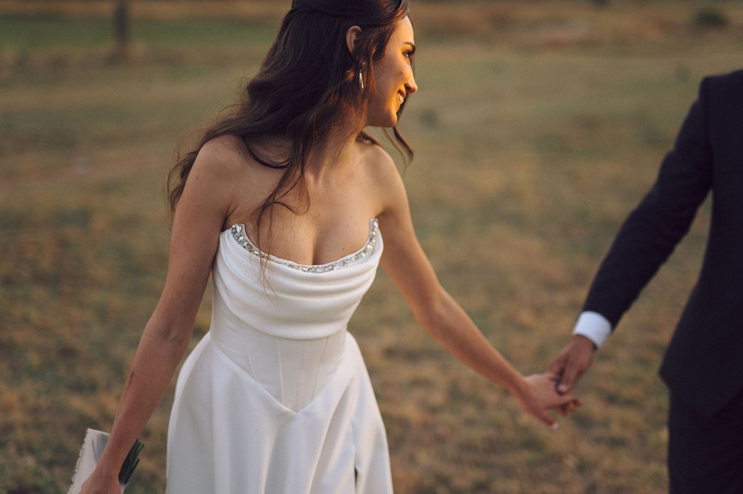Carefree couple dancing together in wedding attire on a grassy field.