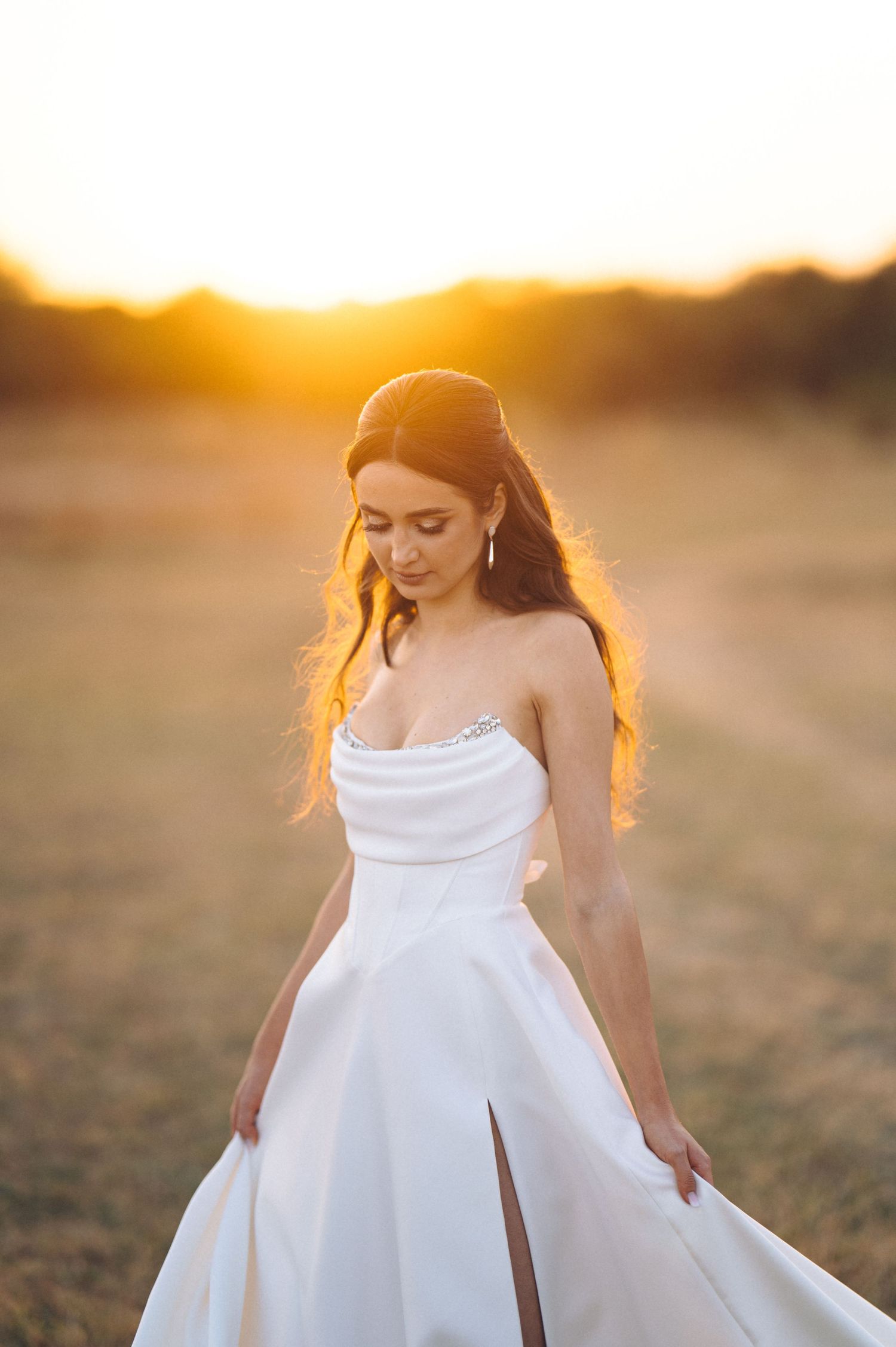 A bride in a flowing white strapless gown stands in sunlit desert landscape with ethereal glow.