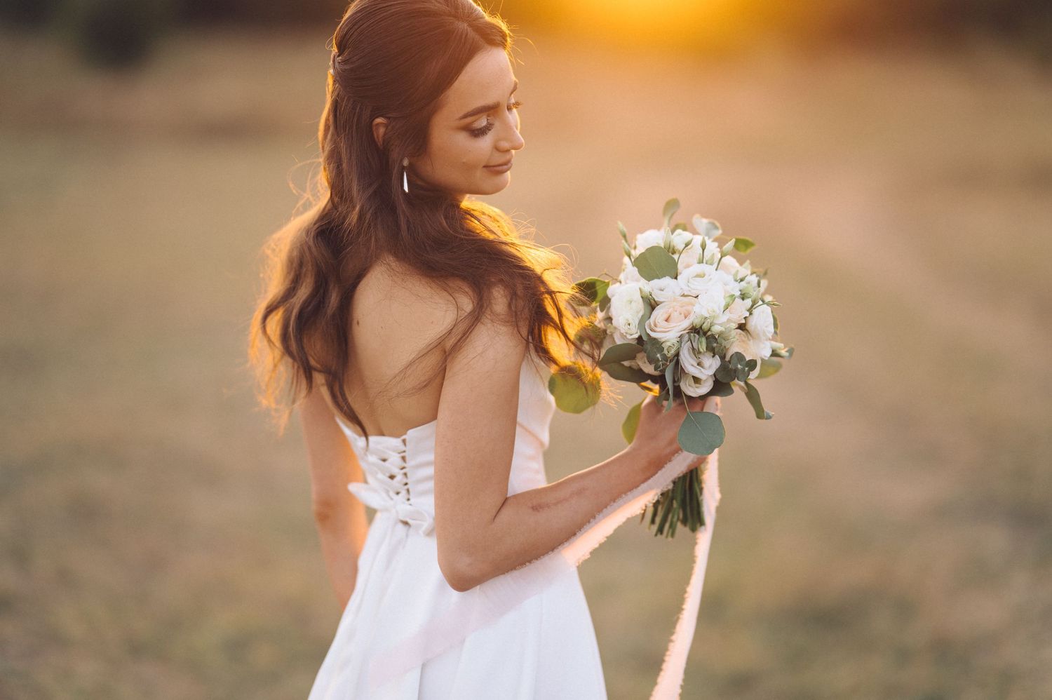 A couple walks along a beach during golden hour sunset holding hands and a white floral bouquet.