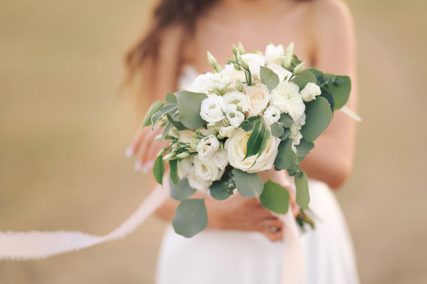 A soft focus white bridal bouquet with eucalyptus leaves and roses held against a blurred cream background.