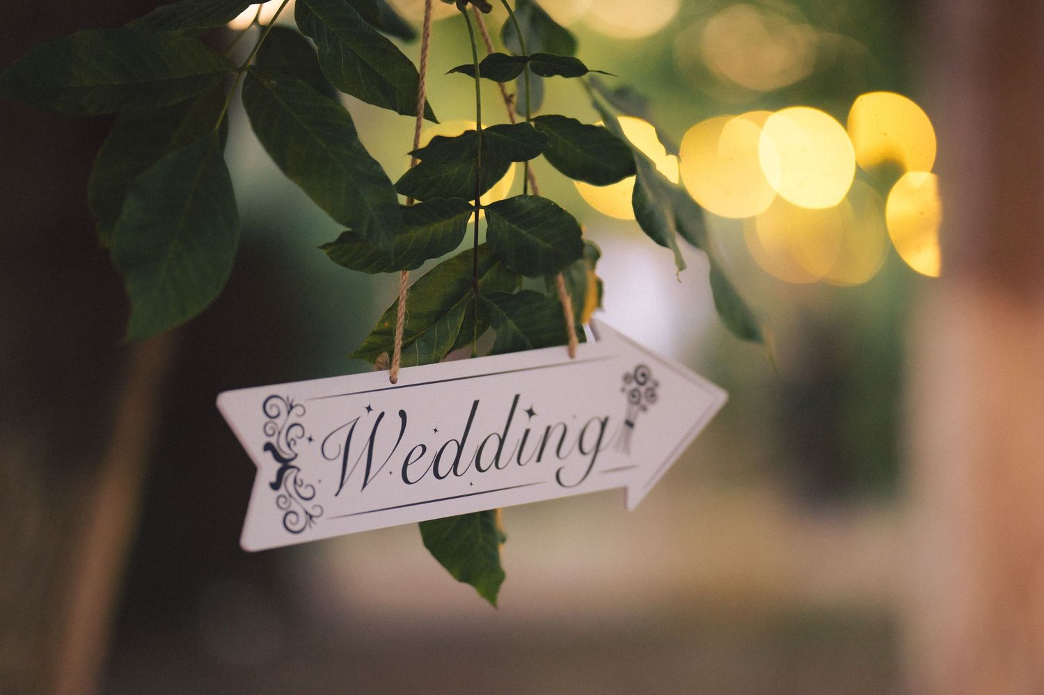 Decorative wedding sign with ornate text hanging amidst blurred bokeh lights and green foliage.