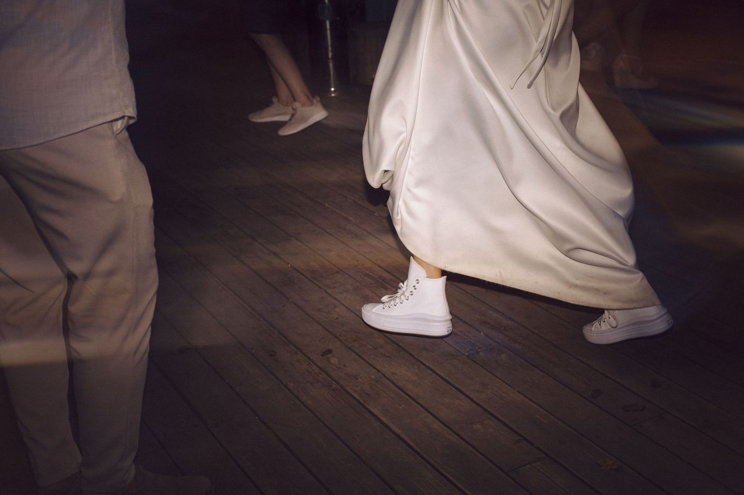 A pair of white sneakers worn with light tan pants walking across a brown wooden floor.