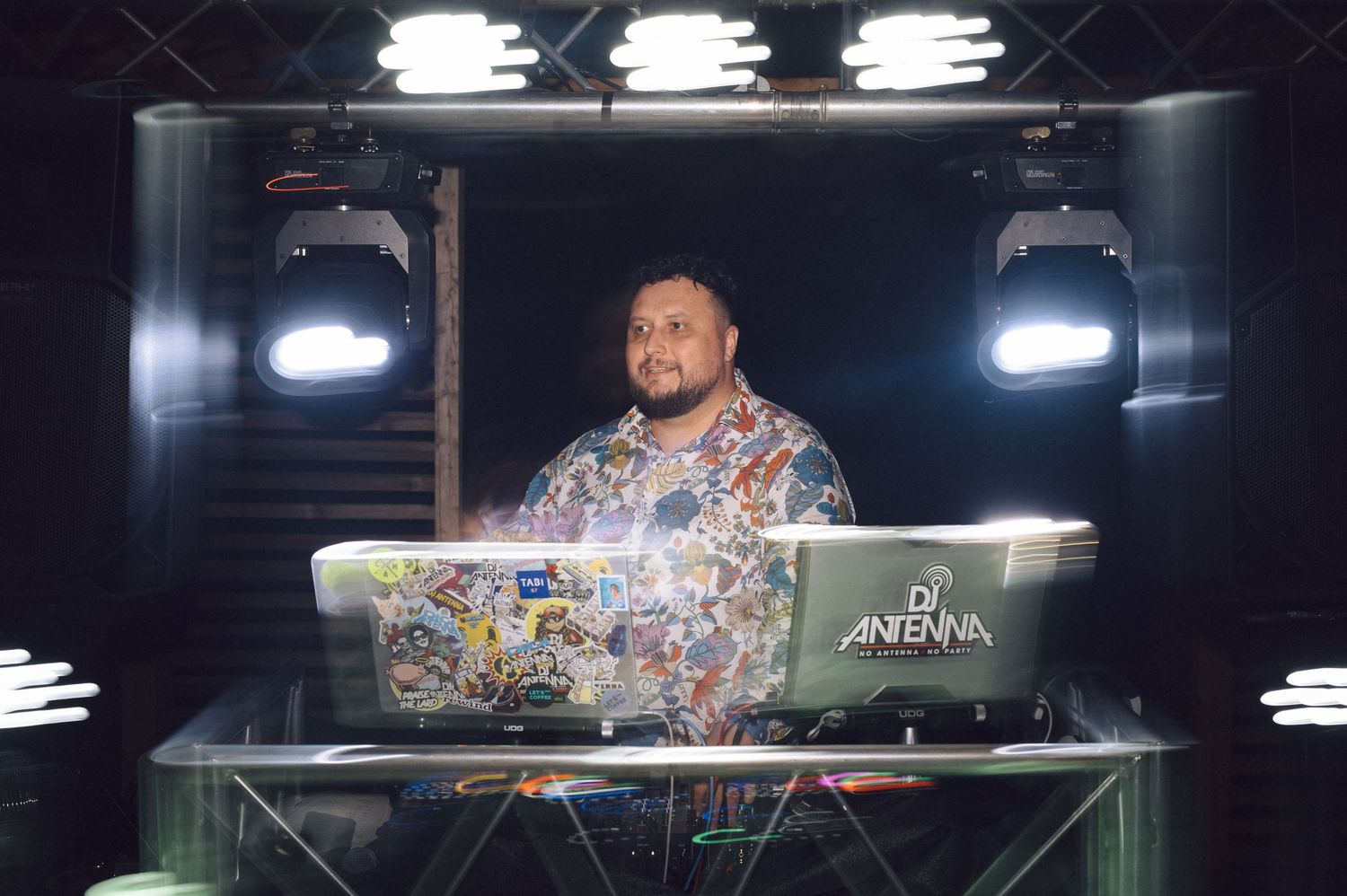 A DJ performs at night behind a glass booth with overhead lighting and stage equipment.