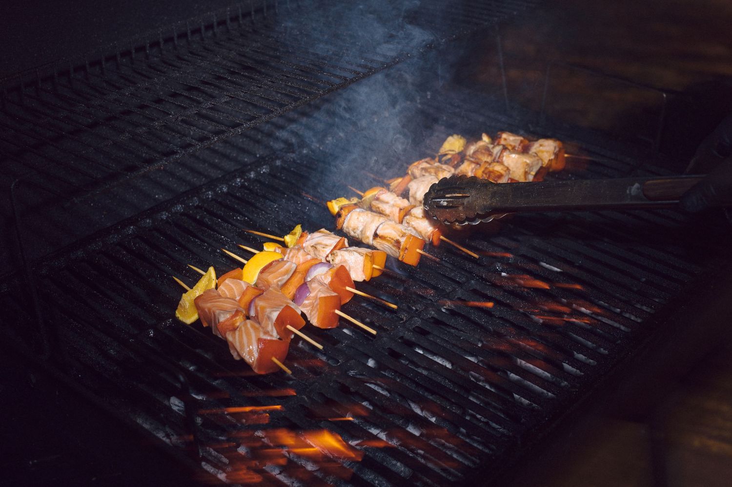 Several kabobs with meat and vegetables cook on a dark grill grate with orange flames visible underneath.