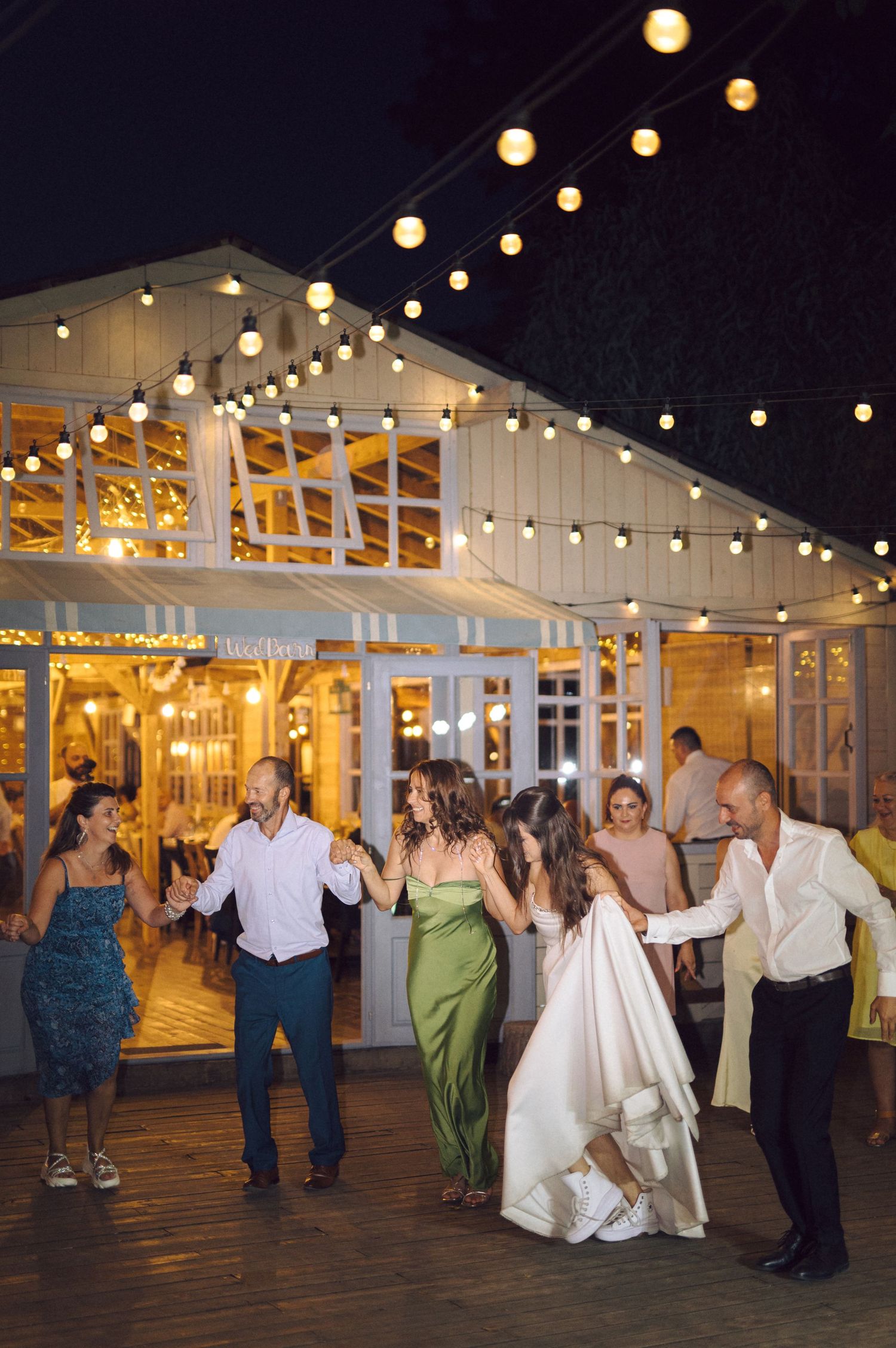 Wedding guests dance under warm glowing string lights at an outdoor evening reception venue.