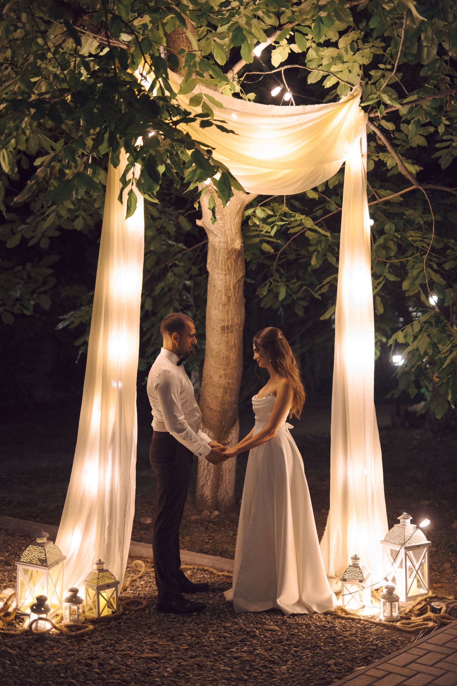 A romantic outdoor wedding ceremony setup with illuminated white fabric draping between trees and lanterns on the ground.