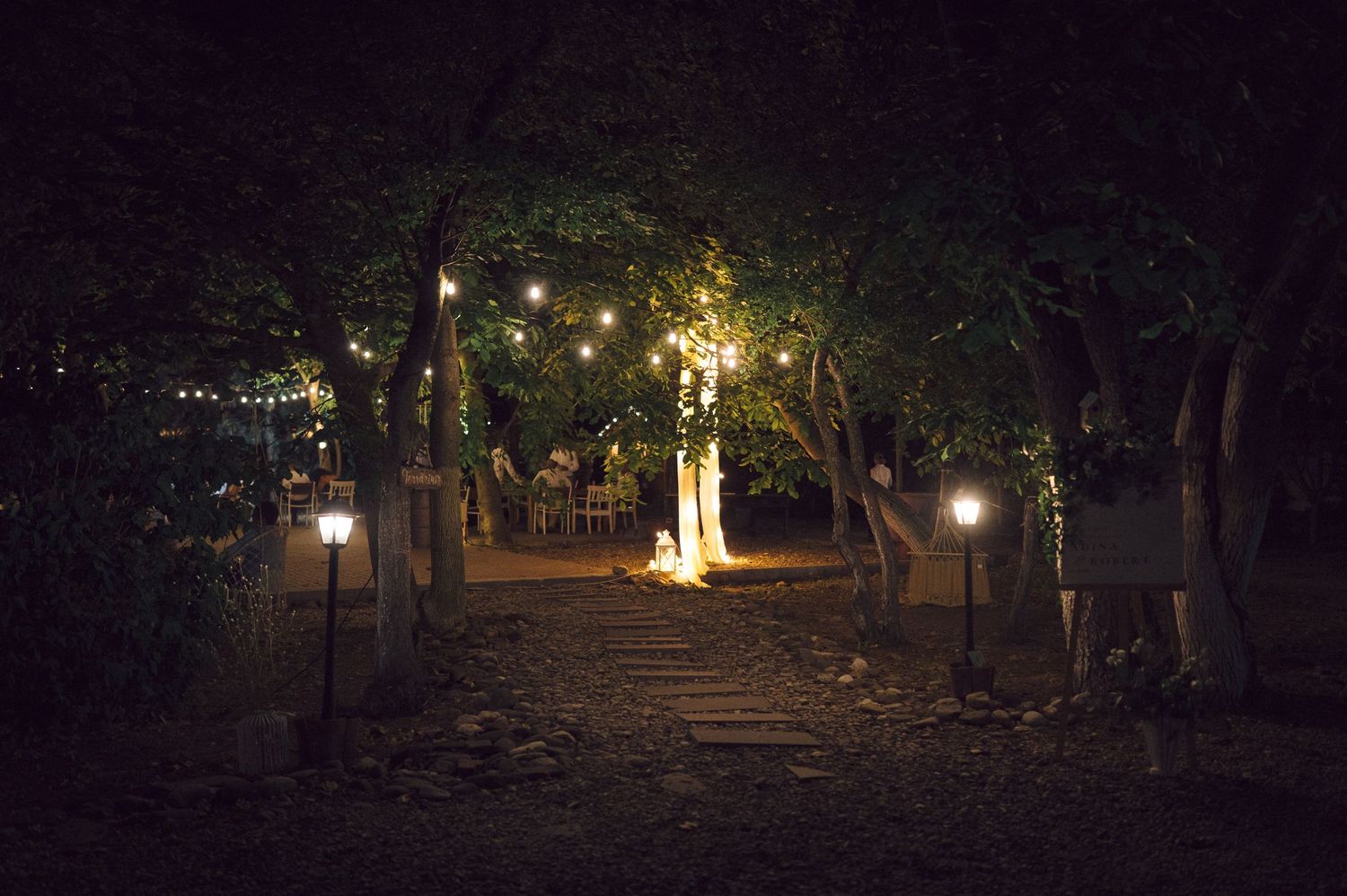 A dimly lit garden path illuminated by warm glowing lanterns and string lights among trees at night.