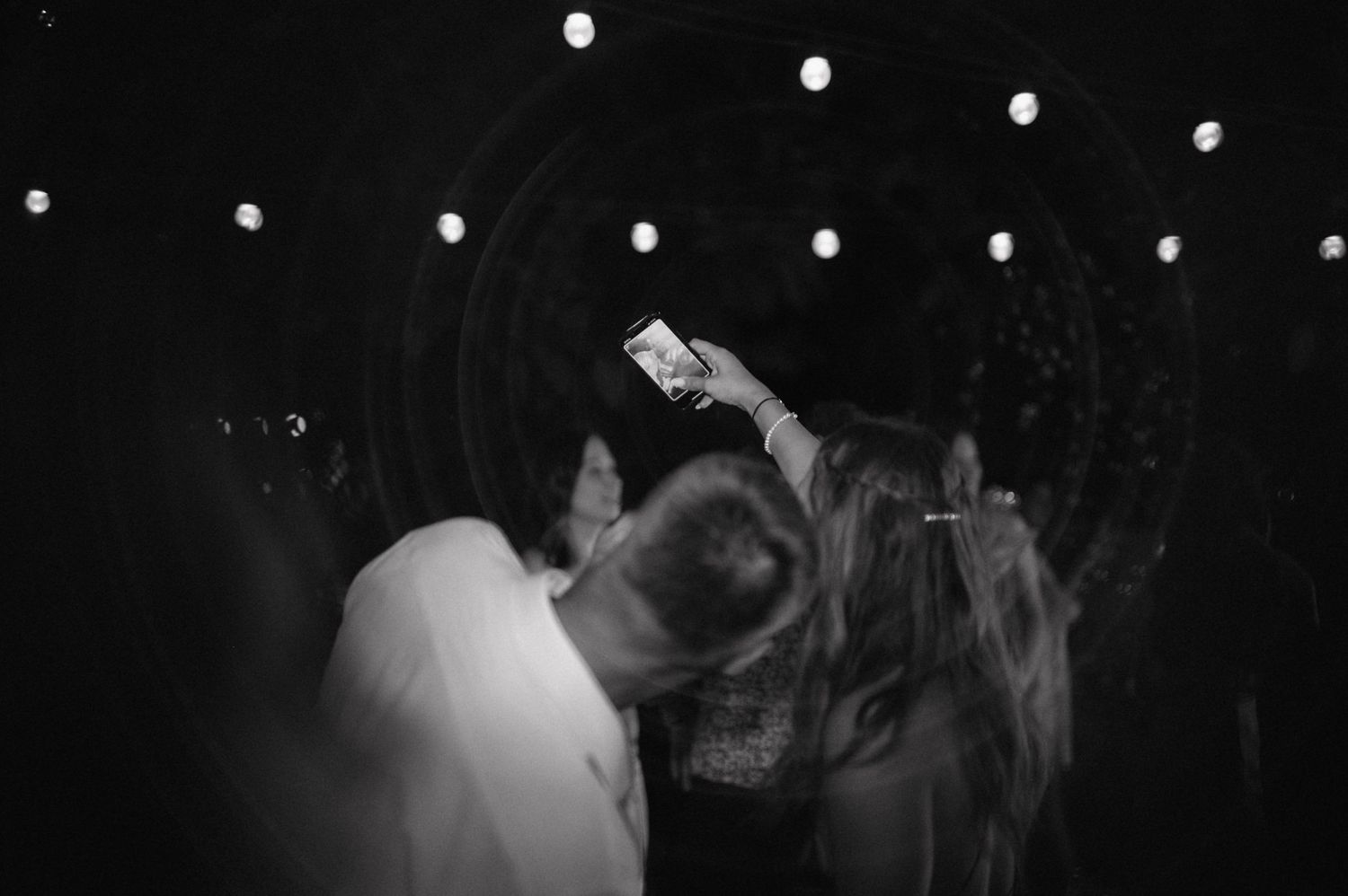 A couple dancing together at a dimly lit event with blurred silhouettes in the background and white lighting overhead.