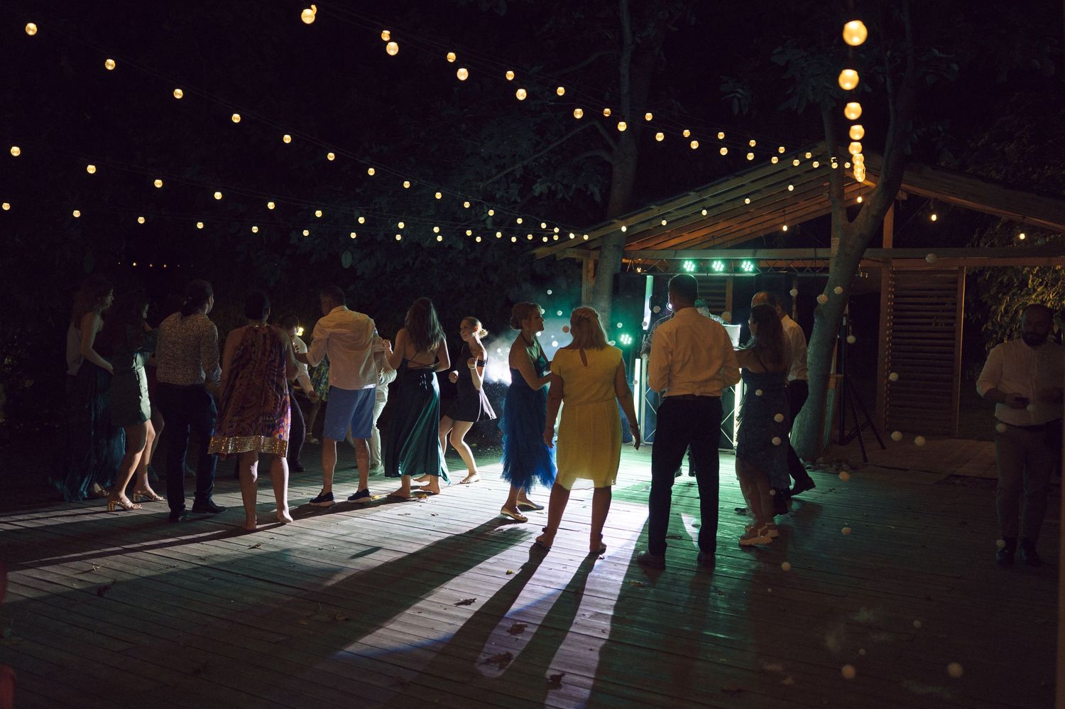 Wedding guests silhouetted against string lights on a dark dance floor at an outdoor evening reception.