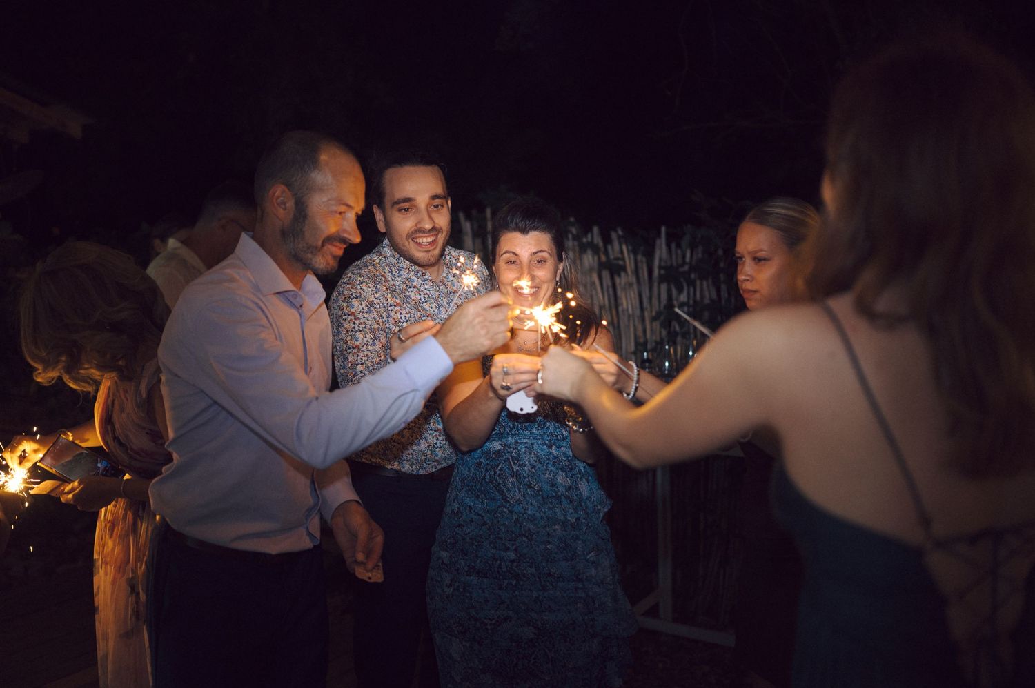 Friends toasting with sparklers and drinks during a nighttime celebration at a dark outdoor venue.