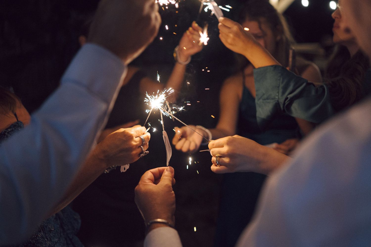 Sparklers illuminate a dark nighttime celebration as partygoers hold them against a black background.