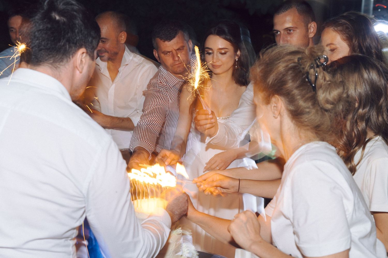 Wedding guests hold sparklers in white attire creating a magical nighttime celebration atmosphere.
