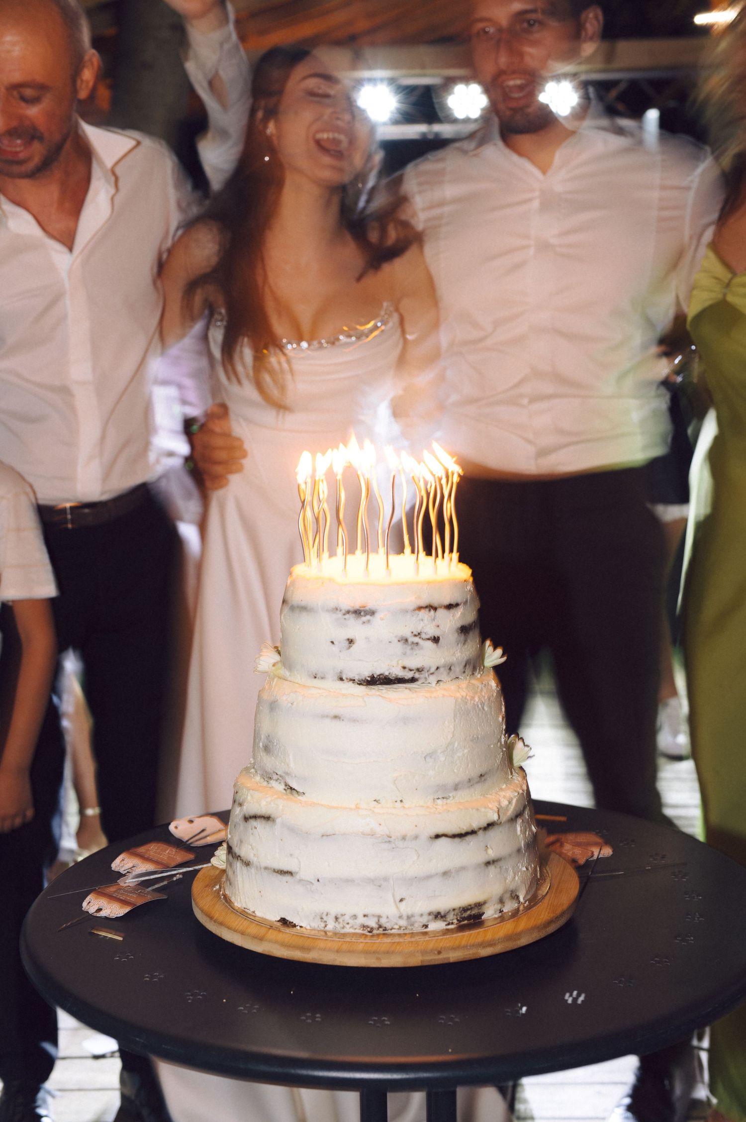 White frosted wedding cake with lit birthday candles glowing brightly on top in a dimly lit reception setting.