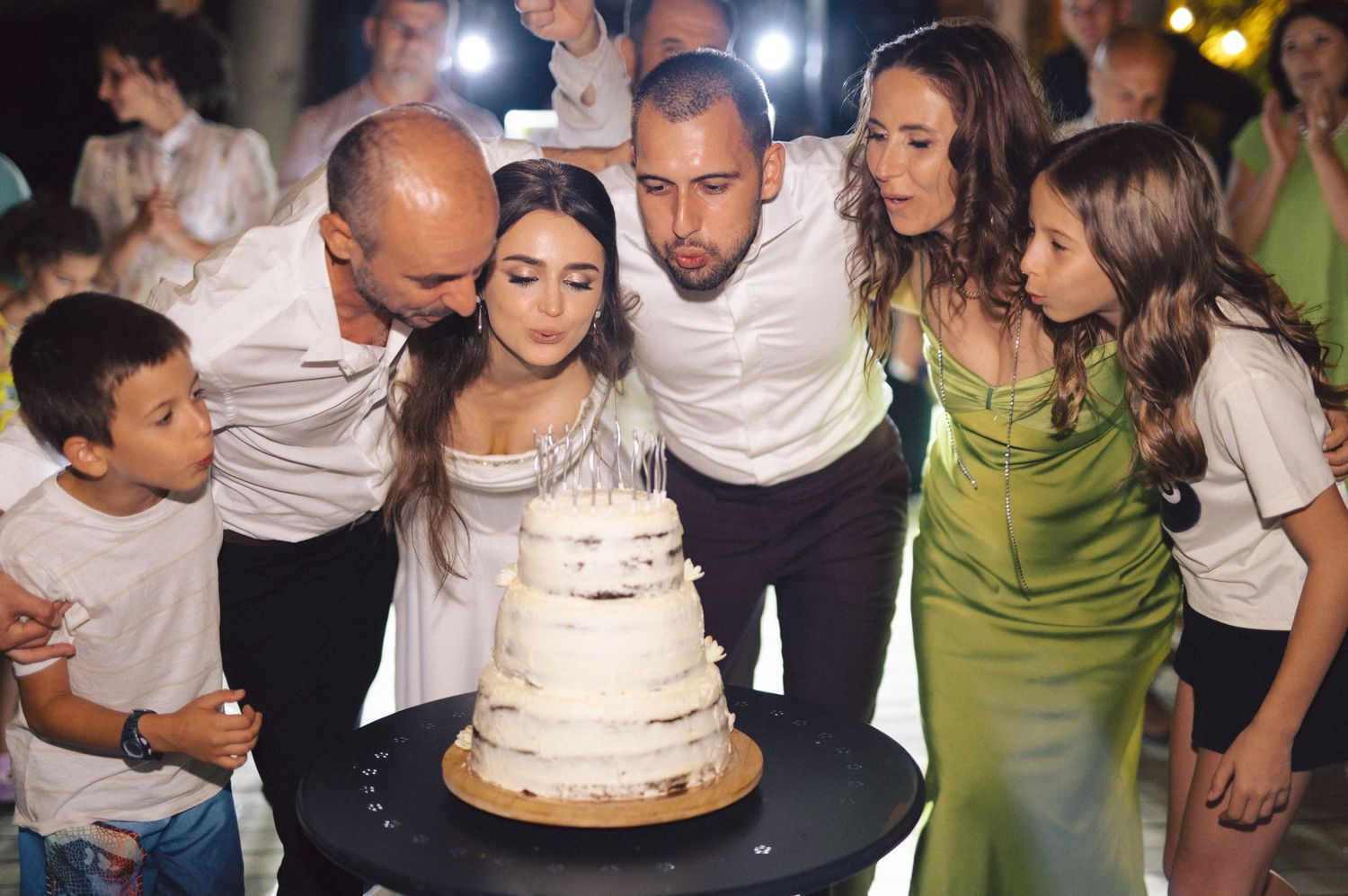A group of party guests lean in together to blow out candles on an elegant white tiered wedding or celebration cake.