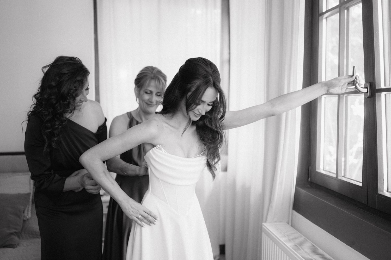 A bride in a white dress gets help with her final preparations near a bright window.