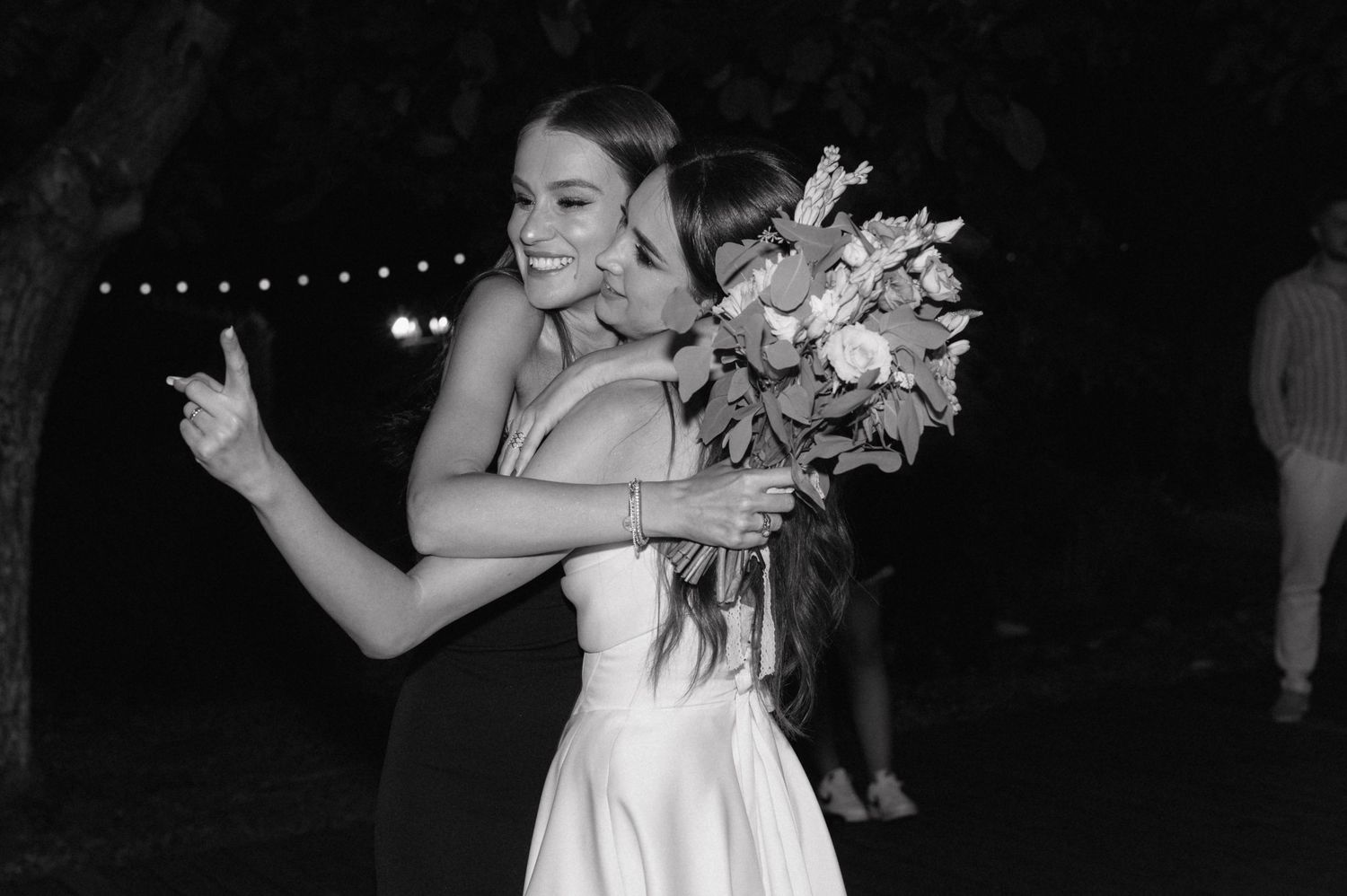 A bride in a white wedding dress dances with a bouquet of flowers at an evening reception with twinkle lights.