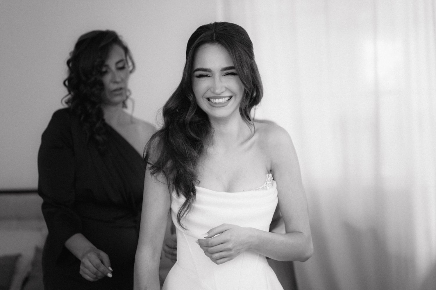 Happy bride in a white dress laughs and smiles with friends during a candid moment before the wedding ceremony.