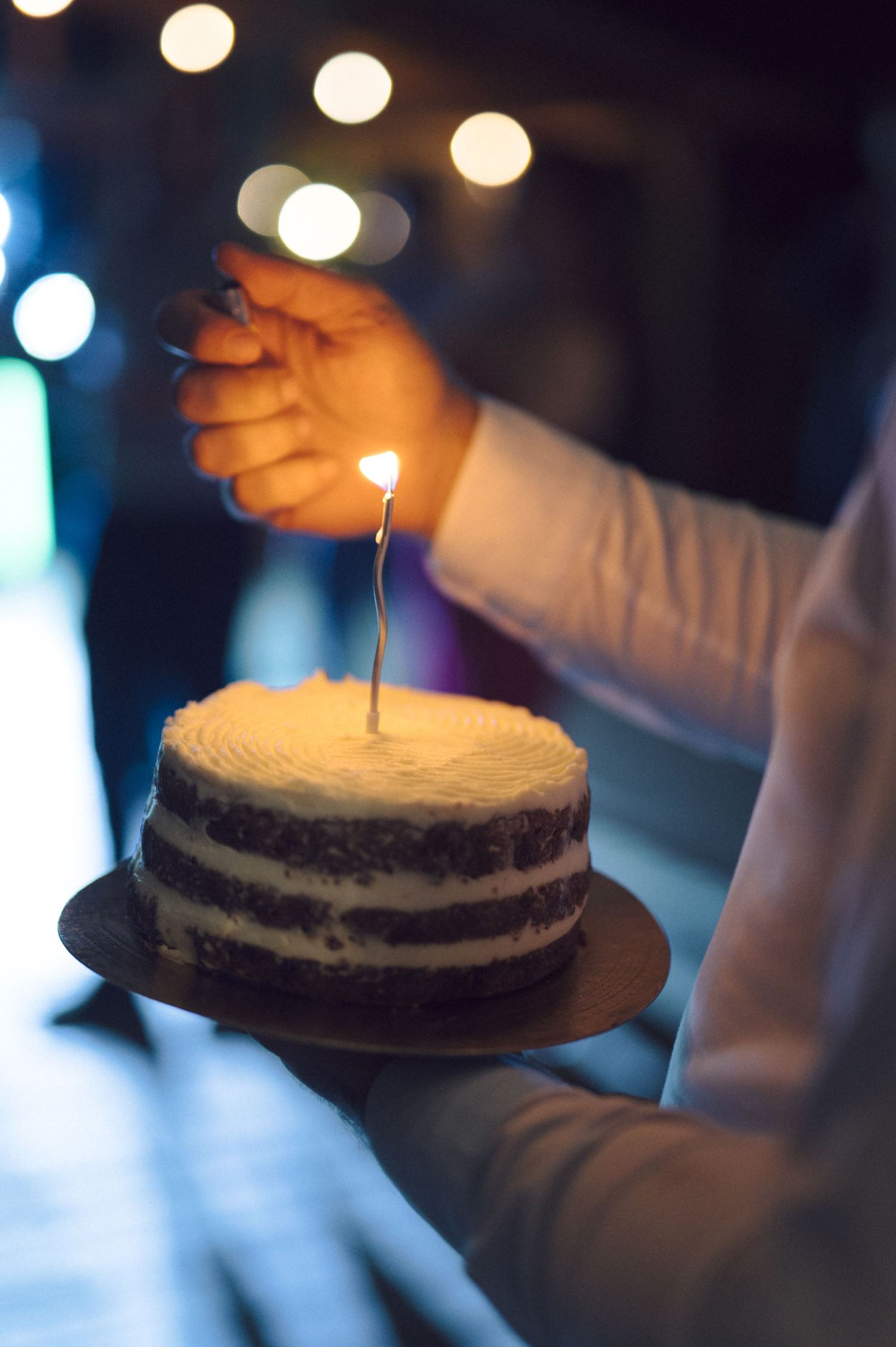 Lighting a birthday candle on a chocolate cream cheesecake in a dimly lit room with bokeh lights in background.