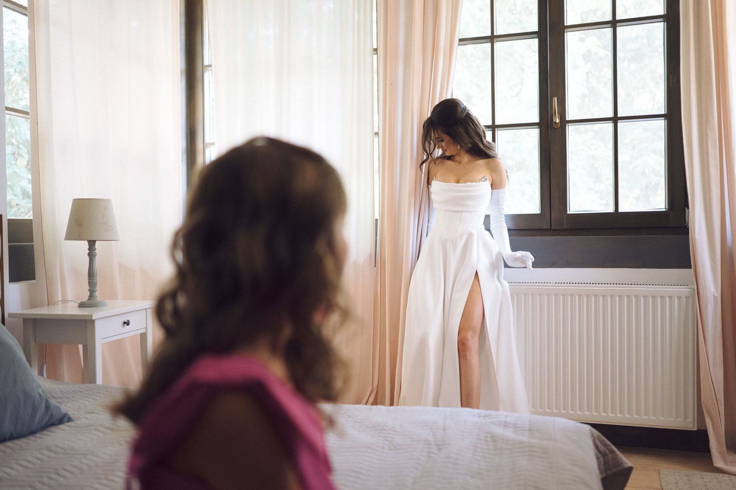 Two people in a bright bedroom with natural light coming through French windows and sheer white curtains.
