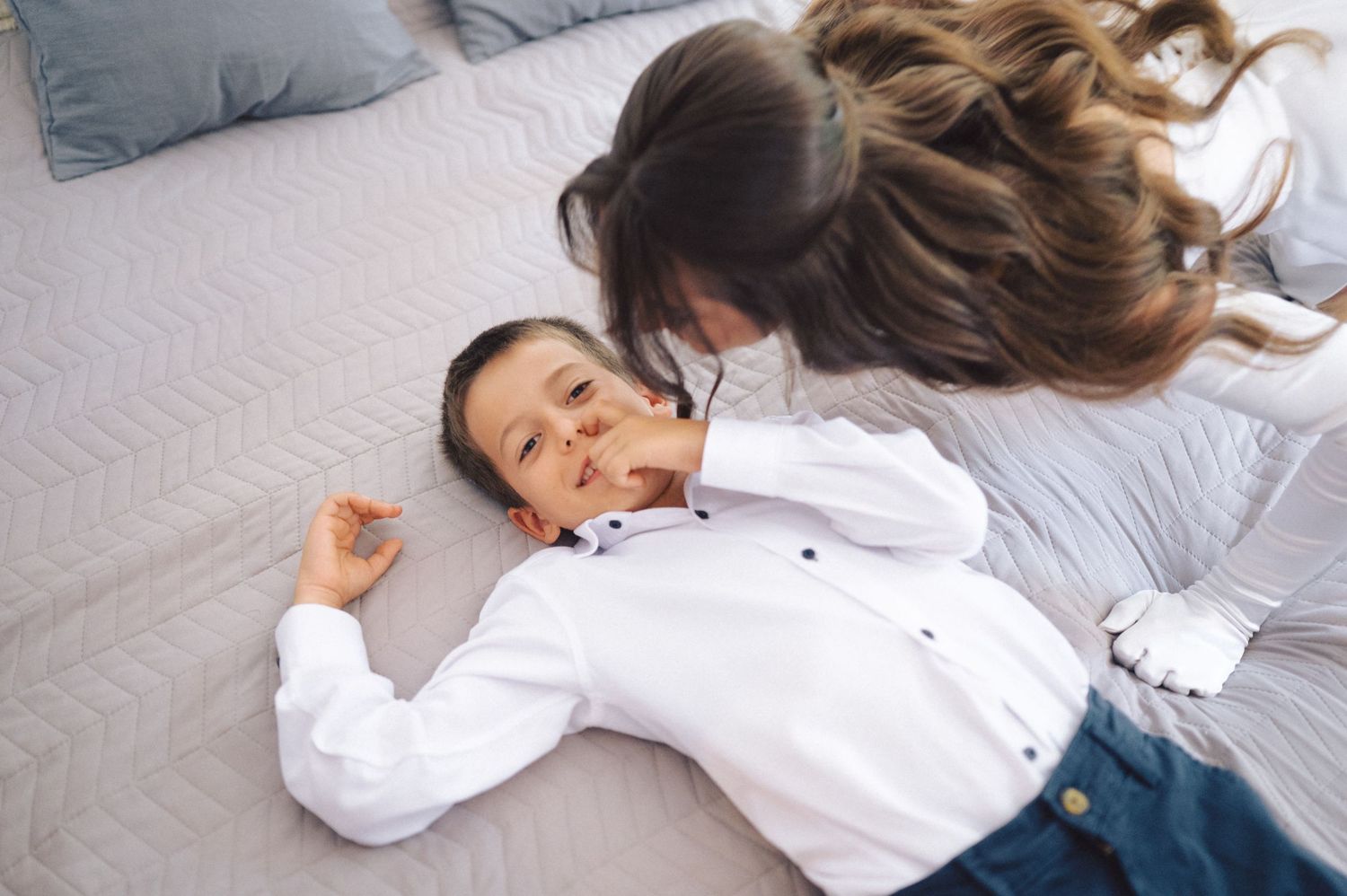 A playful child wearing a white shirt rolls and poses on cozy grey bedding in a bedroom.