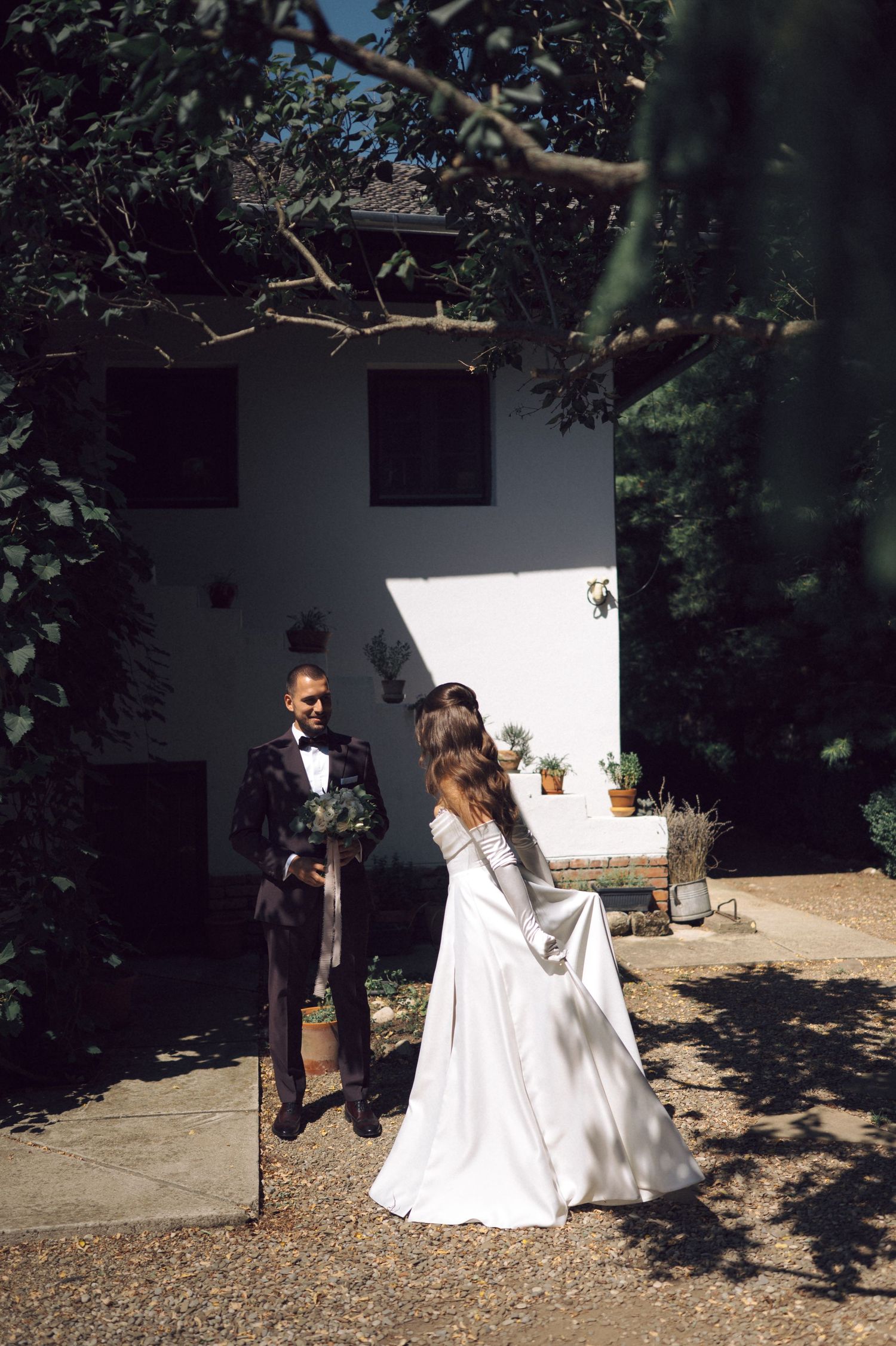 A couple in wedding attire walks together on a sun-dappled outdoor path with white walls and greenery in the background.