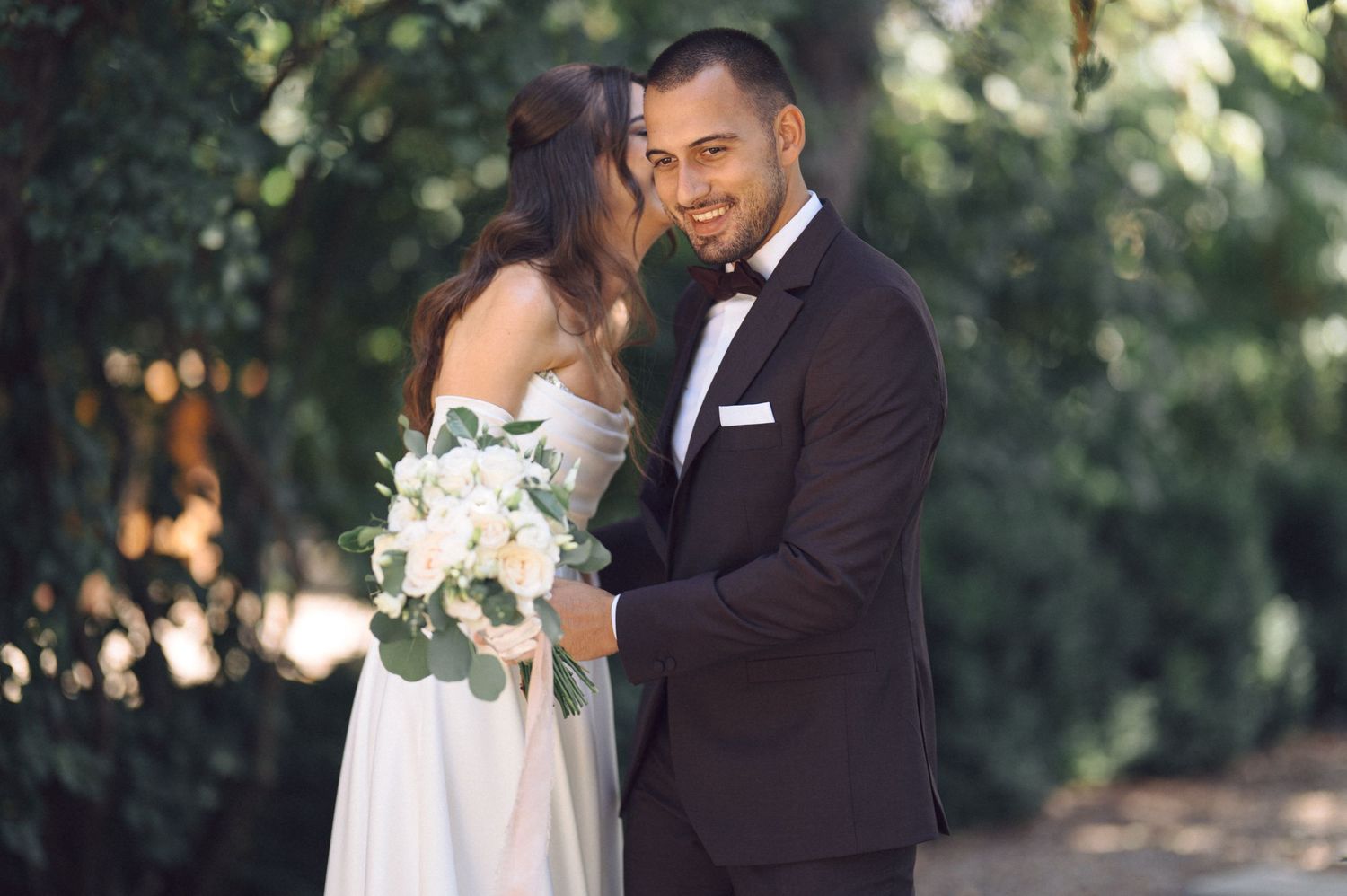 A romantic outdoor wedding photo shows a couple embracing among lush greenery while the bride holds a white floral bouquet.