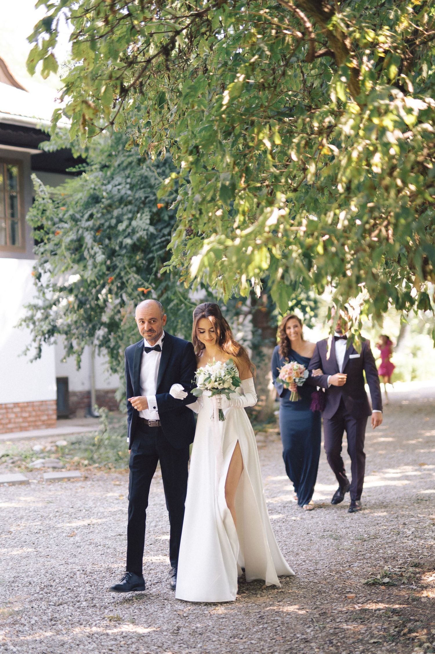 Wedding party members walking together down a tree-lined path in formal attire.