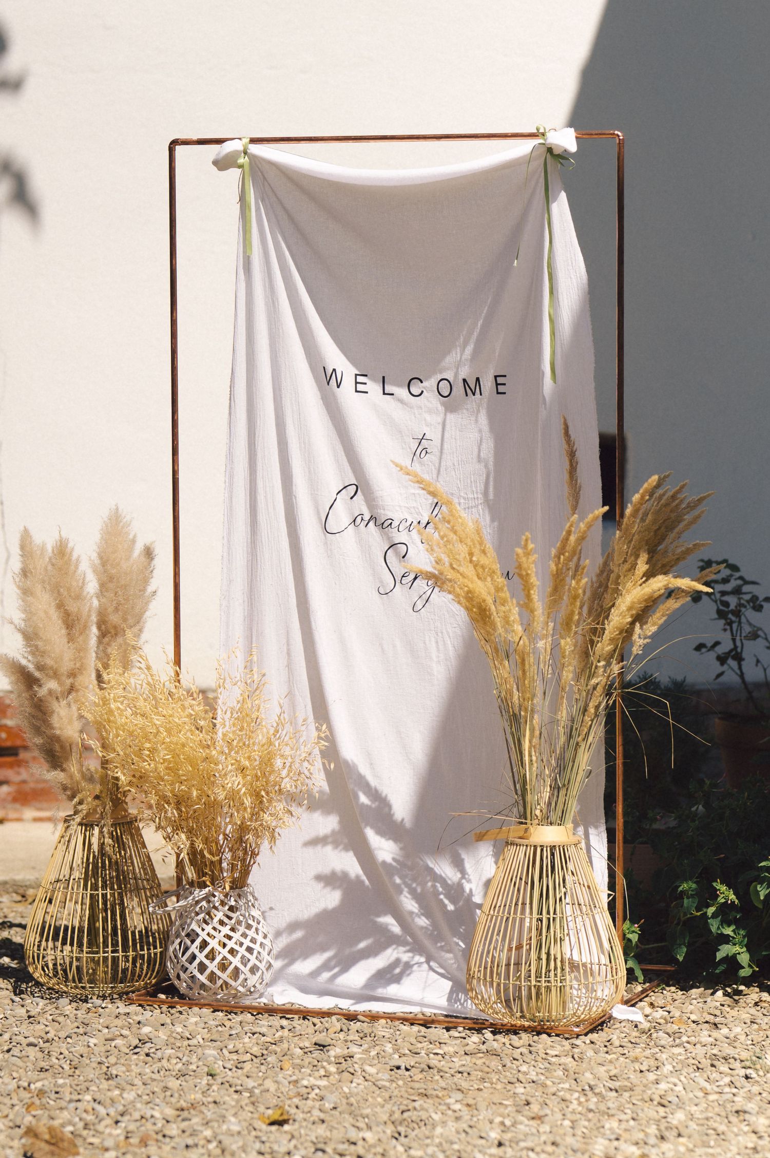 A white welcome banner hangs on a copper frame with dried pampas grass and wicker vases displayed at the base.