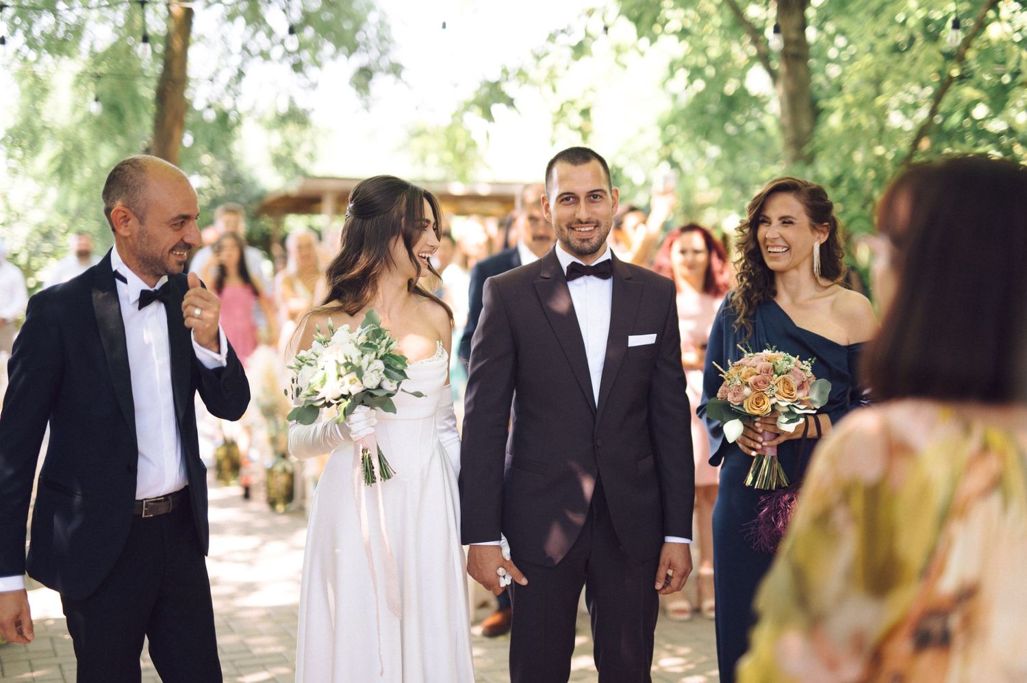 Wedding celebration outdoors with guests in formal attire gathering for festivities in a natural garden setting.