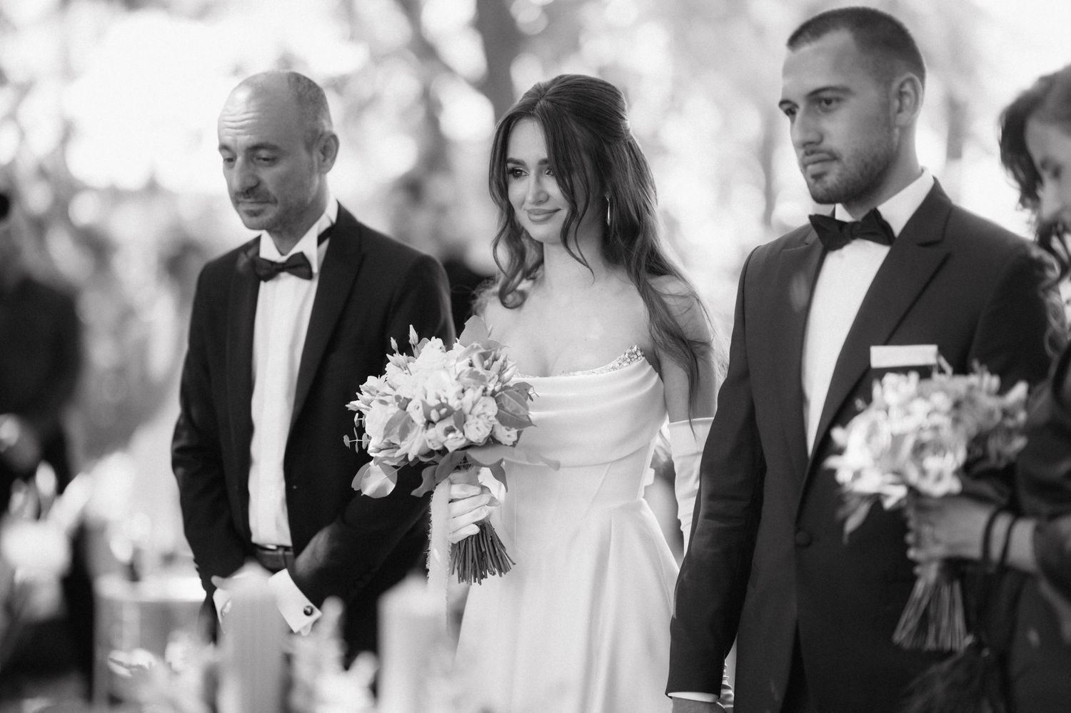 Black and white wedding ceremony photo of wedding party members in formal attire holding floral bouquets.