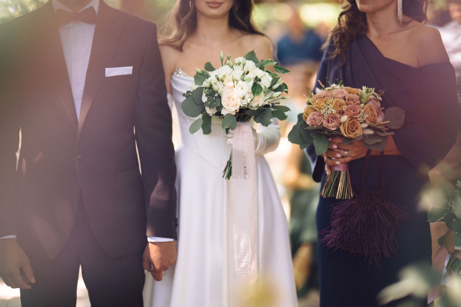 Elegantly dressed guests at a formal outdoor wedding event carry champagne and floral bouquets.