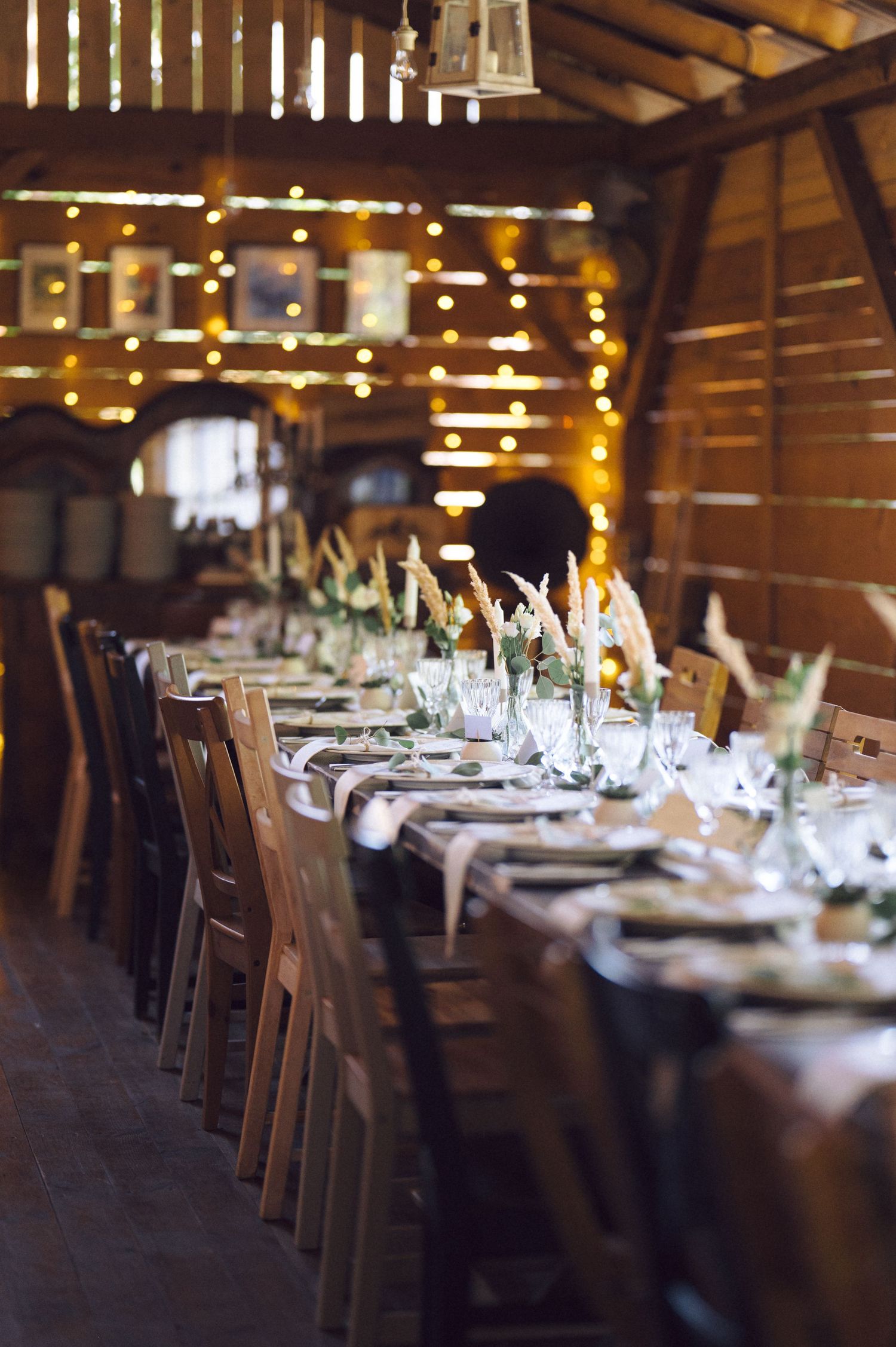Elegant rustic wedding reception setup in a wooden barn with long tables, white tablecloths and twinkle lights overhead.
