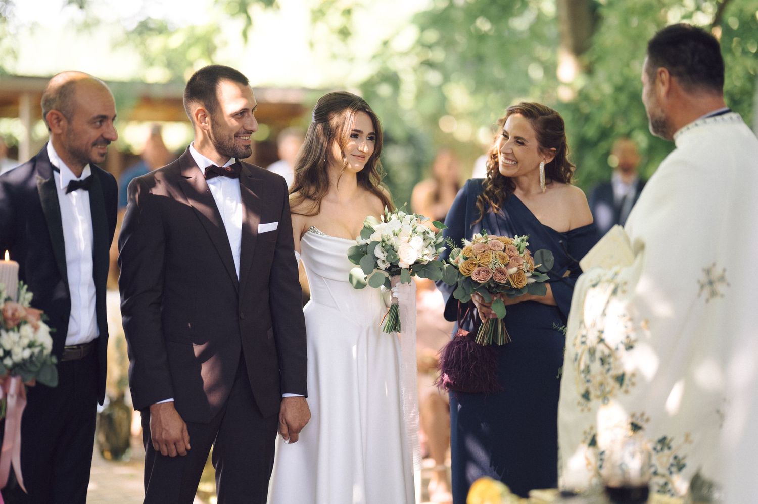 Guests and wedding party celebrate at an elegant outdoor garden reception with sunlit trees in the background.