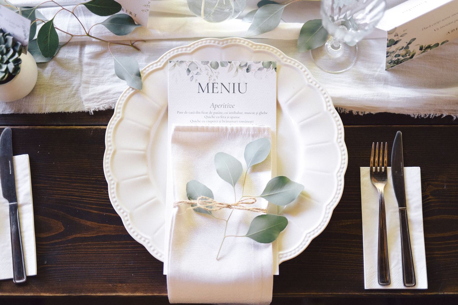 Elegant place setting with white plate, menu card, eucalyptus leaves and silverware on dark wooden table.