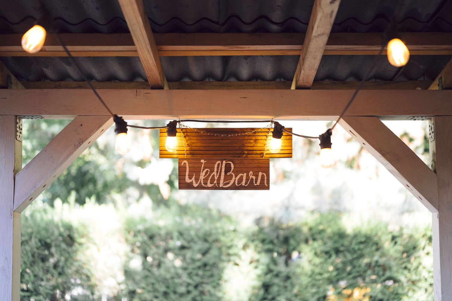 String lights illuminate a rustic barn's overhang with a wooden sign and lush greenery peeking through.