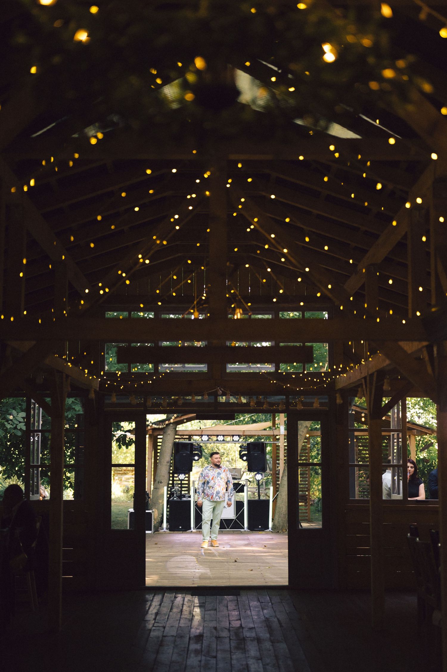 Christmas lights create a magical ambiance along a covered wooden walkway leading to a bustling outdoor pavilion.