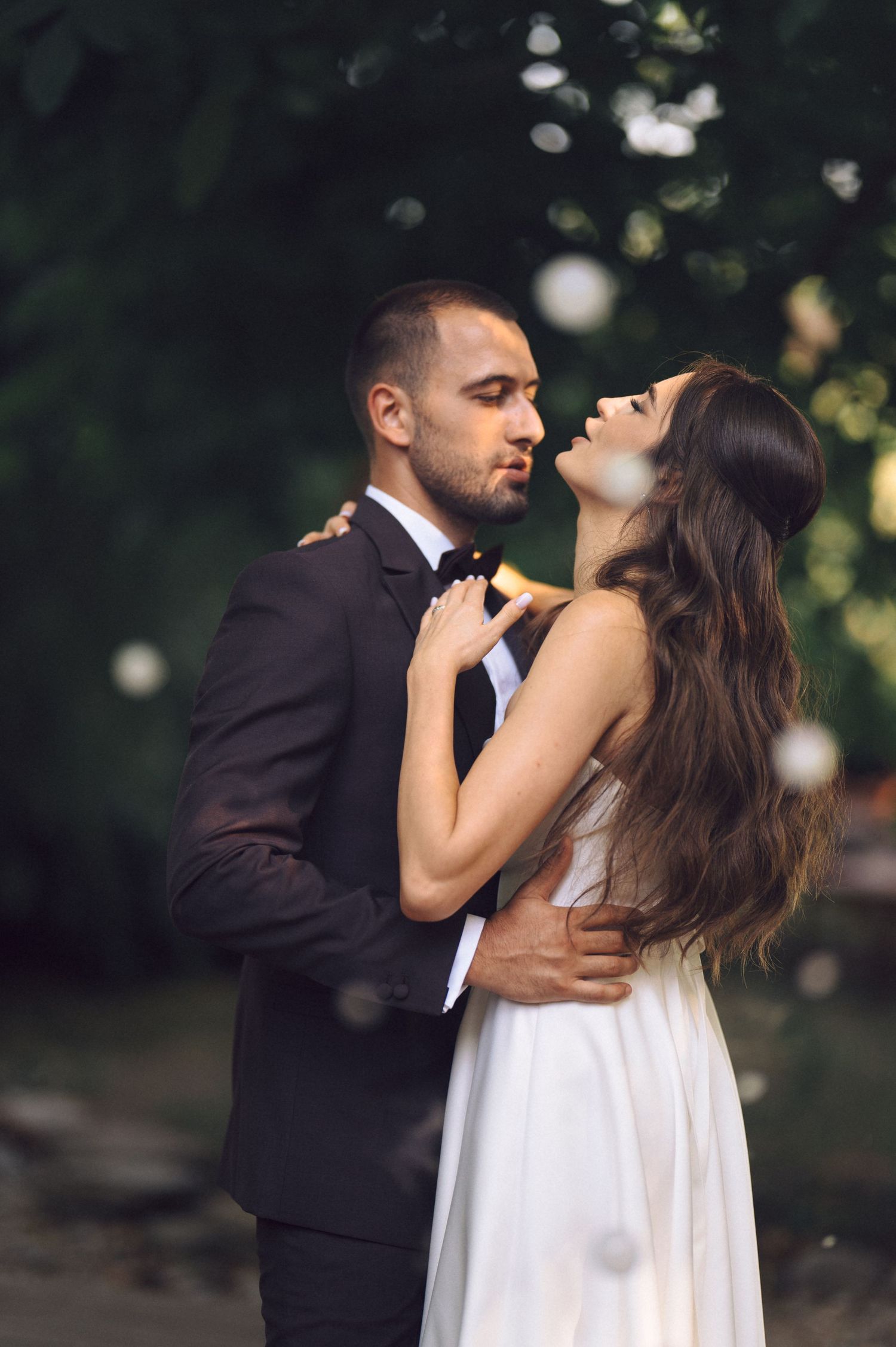A couple in wedding attire share a tender outdoor embrace under soft bokeh lighting.
