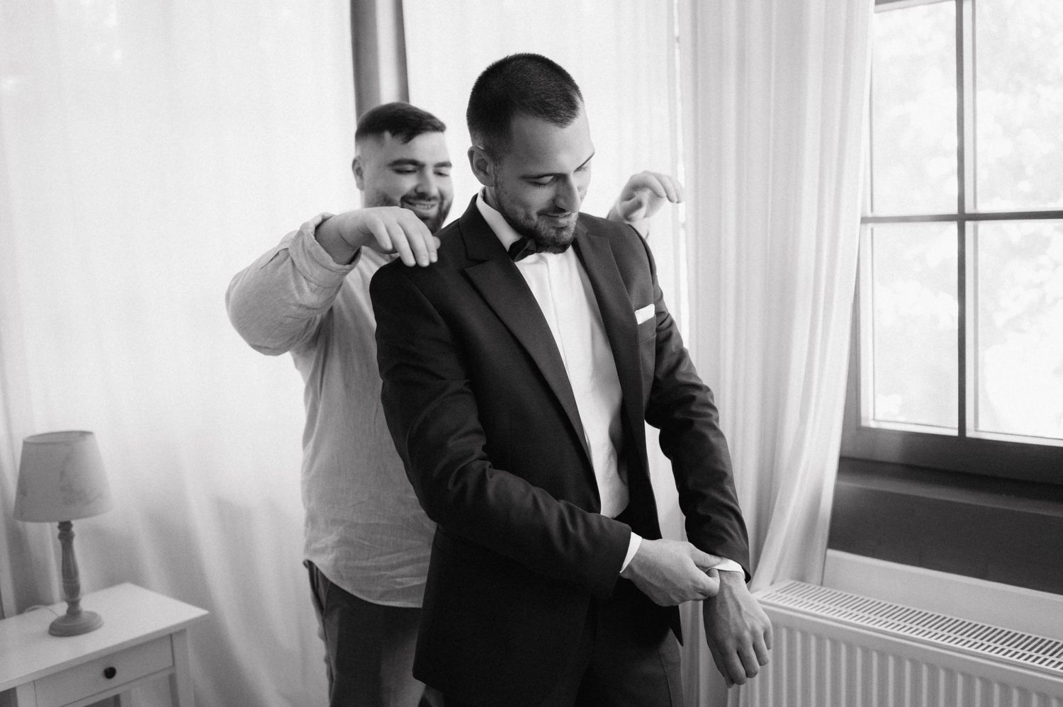 Groom and groomsmen get ready for wedding in elegant black tuxedos while standing near sunlit window.
