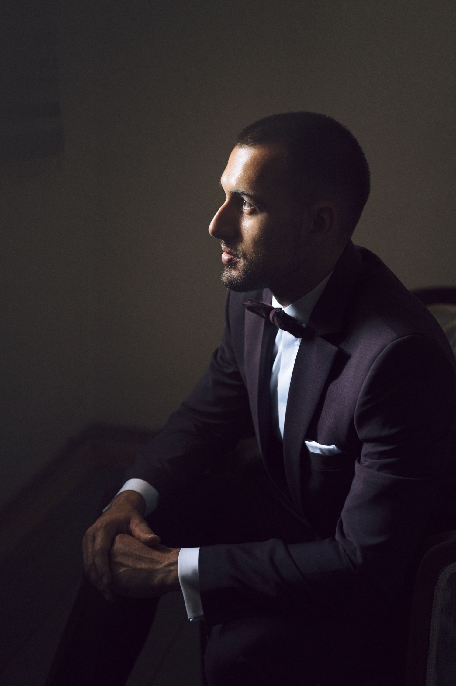 A person in a black tuxedo sits thoughtfully in dramatic low-key lighting against a dark background creating a moody portrait.