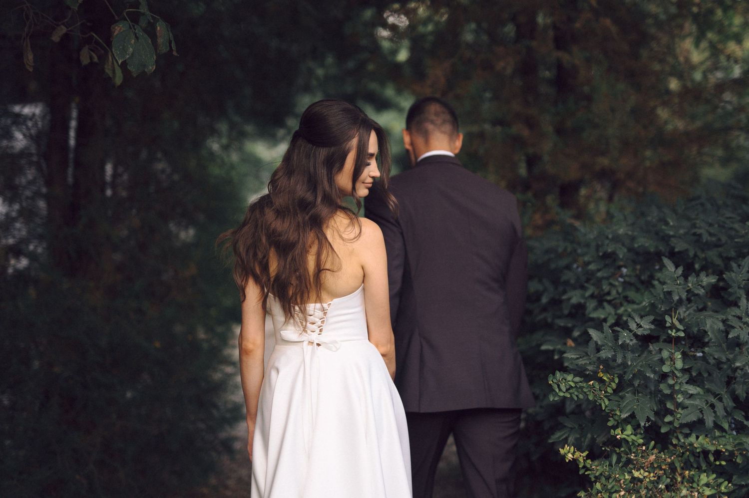 Wedding couple strolls along garden path surrounded by green foliage.